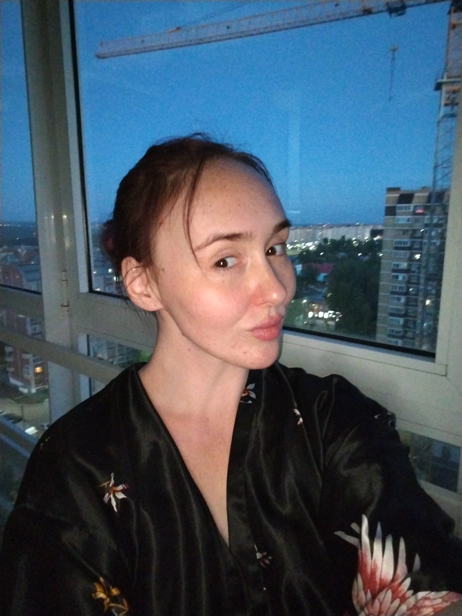 A woman is featured in a close-up portrait, seemingly a selfie, taken indoors next to a large window. She looks directly at the camera with a playful expression, her lips pursed as if blowing a kiss or pouting. Her hair, which is dark brown, is pulled back from her face, revealing her forehead and freckles across her cheeks and nose.
She is dressed in a dark, possibly black, silky or satin robe adorned with prominent white and red floral or bird-like patterns on the shoulder and lower front, along with subtle golden accents elsewhere.
The background through the window displays an urban landscape at dusk or twilight. The sky is a deep blue, and lights from various buildings and streets illuminate the city below. A large construction crane stands tall against the sky on the upper right, indicative of urban development. Below, several multi-story buildings and areas of trees are visible, extending into the distance. The view suggests the image was taken from a high vantage point, such as an apartment building in Izhevsk, Russia. No visible text is present in the image.