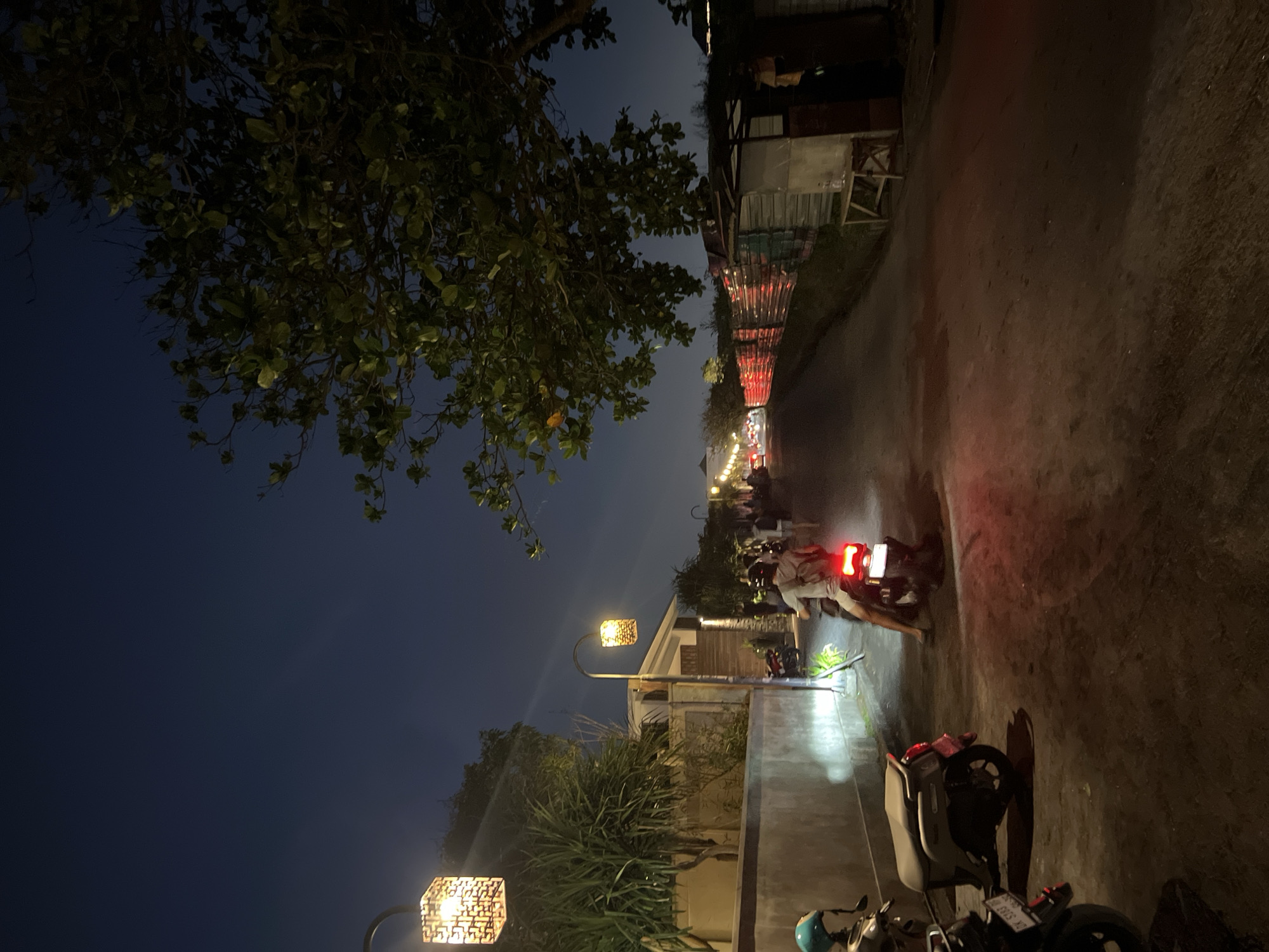 This nighttime shot captures a narrow, seemingly paved road lined with structures on either side. The image is taken in the city of Canggu, Indonesia. Two motorcycles are visible: one on the right side of the road, partially cropped, and another further down the road being driven away from the camera. The street is illuminated by decorative street lamps, and red lights are visible further along the road, possibly indicating more vehicles or decorations. A large tree is positioned on the left side of the image, its leaves partially obscuring the sky. The sky appears dark blue to black, indicating a late evening or night setting.