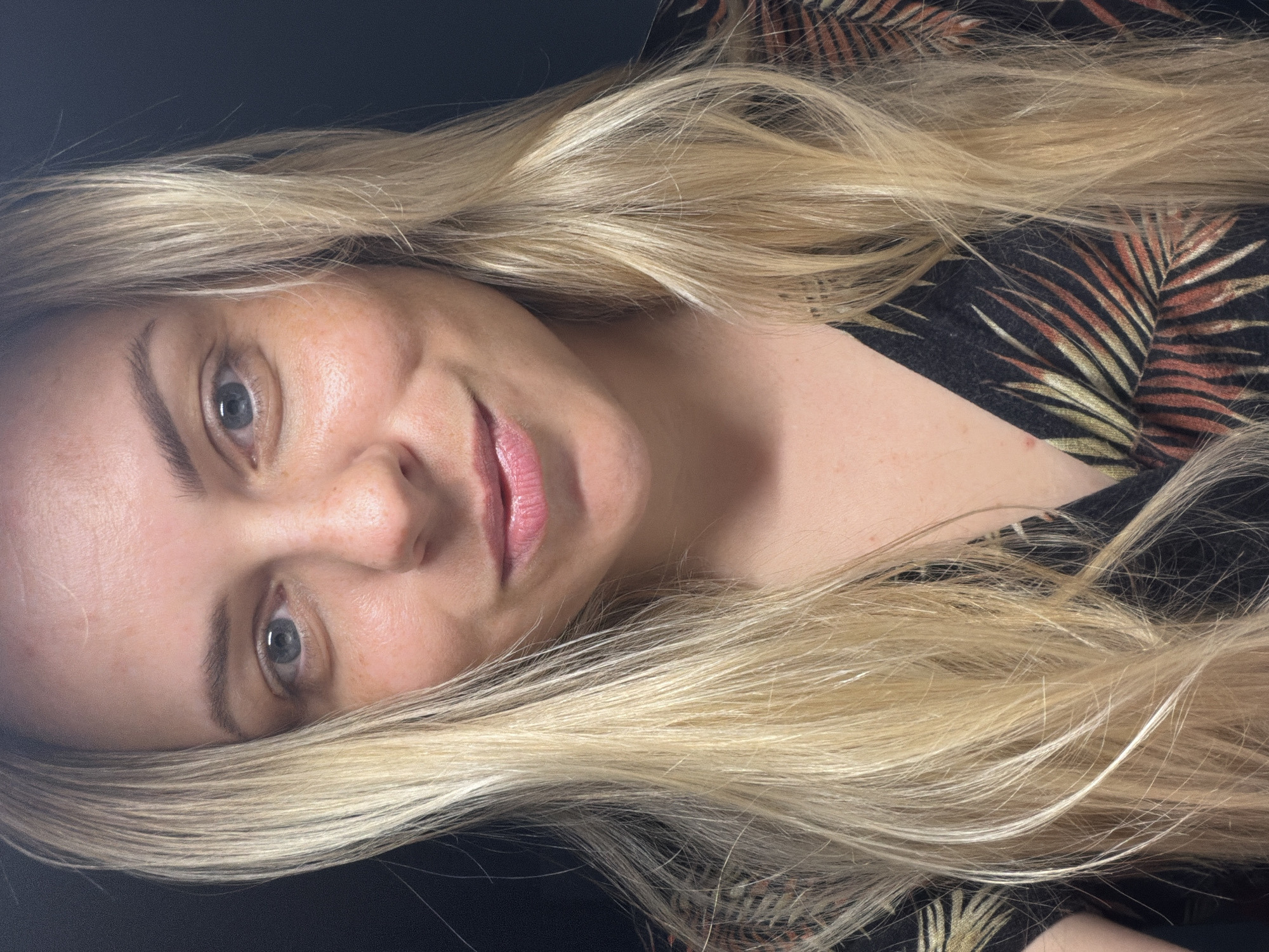 A close-up, head-and-shoulders portrait of a blonde woman with blue eyes and fair skin. She is looking directly at the camera with a slight smile. Her hair is long and wavy, falling around her shoulders. She is wearing a dark top with a tropical leaf pattern. The background is dark and out of focus. There is no discernible location in Votkinsk, Russia.