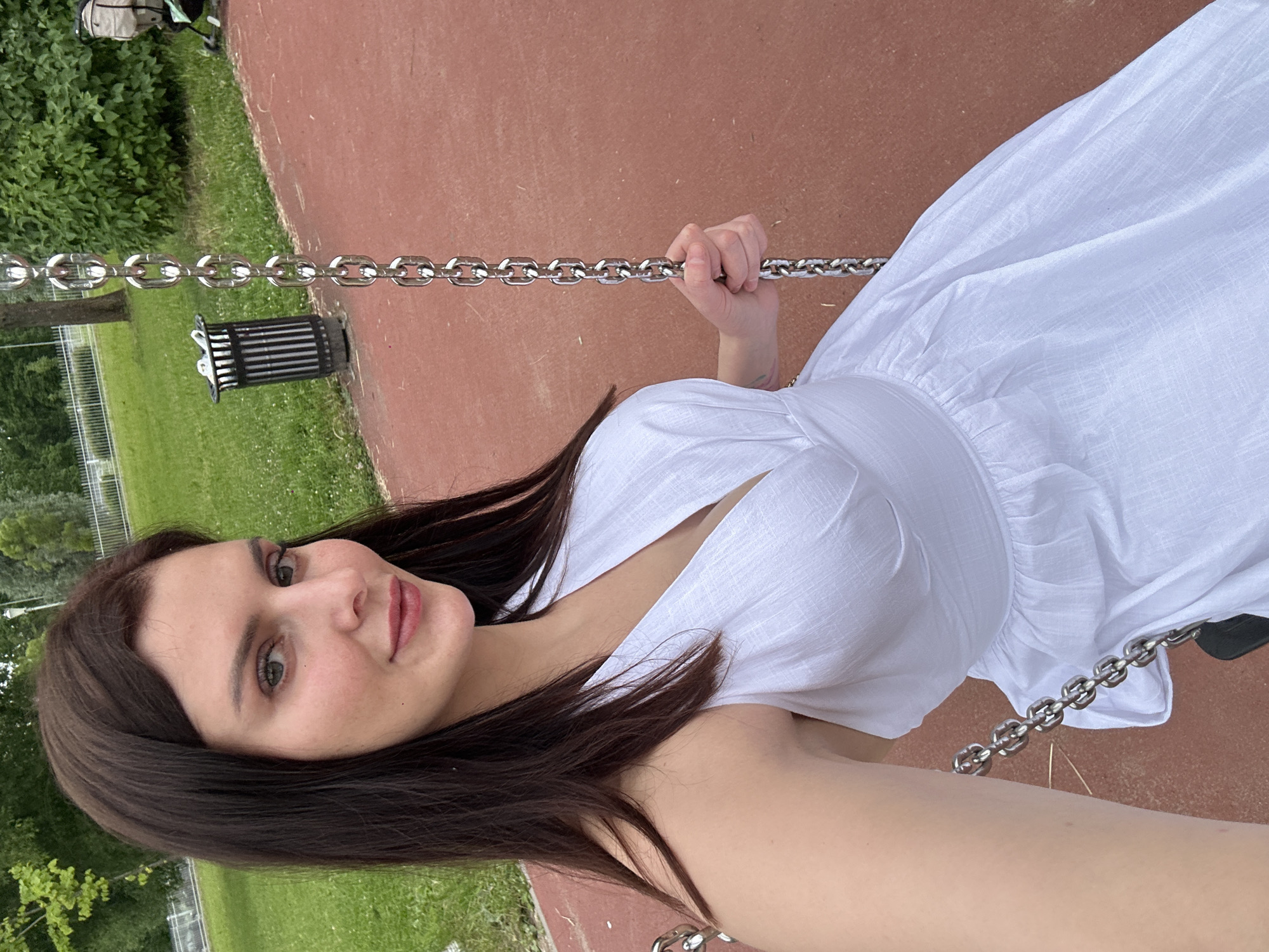 A medium shot shows a woman with long, dark brown hair and light skin, wearing a white, sleeveless dress with a V-neck and ruffles, as she sits on a swing in an outdoor park. The swing chains are made of thick, silver metal. The woman's right arm is visible, holding onto the chain. Her skin is smooth and her makeup is subtle, with rouged cheeks and pink lipstick. She is looking towards the camera with a gentle smile. The background shows a grassy area with trees and bushes, a black trash can with silver accents, and a red surface that appears to be a playground floor. A stroller is visible in the far right background.