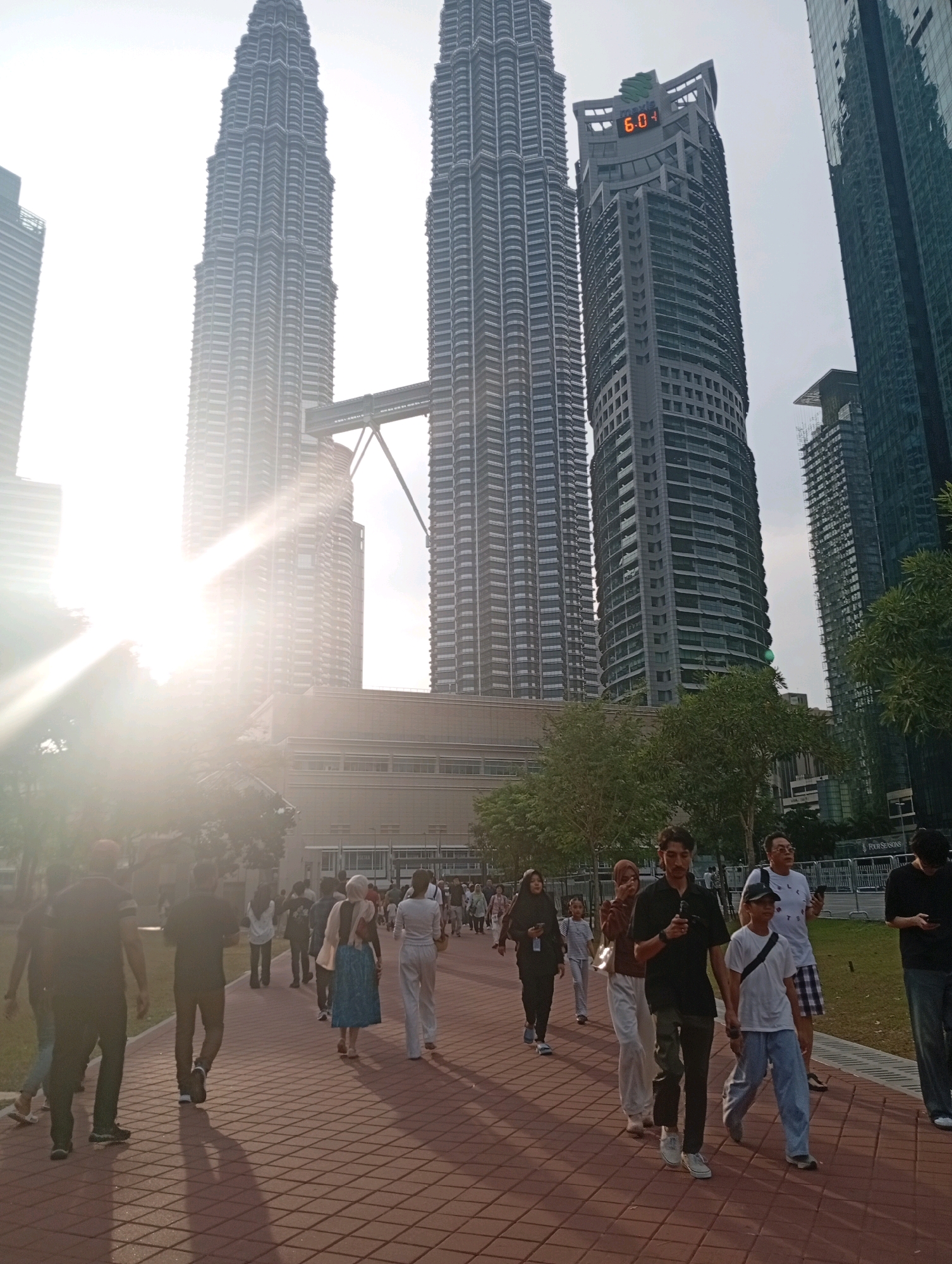 This image captures a view of the Petronas Twin Towers in Kuala Lumpur, Malaysia, on a bright, sunny day with a strong lens flare effect. A group of people are walking on a brick pathway in the foreground. The people are diverse in age and attire, with some wearing casual clothing and others headscarves. The pathway is paved with reddish-brown bricks and is lined with trees and grass to the right. In the background, the iconic Petronas Twin Towers dominate the skyline, connected by a skybridge. To the right of the towers, another tall skyscraper is visible, displaying a digital clock showing "6:04". The sky is bright and somewhat hazy, indicating daytime. The overall scene suggests a tourist or public area in front of the landmark.