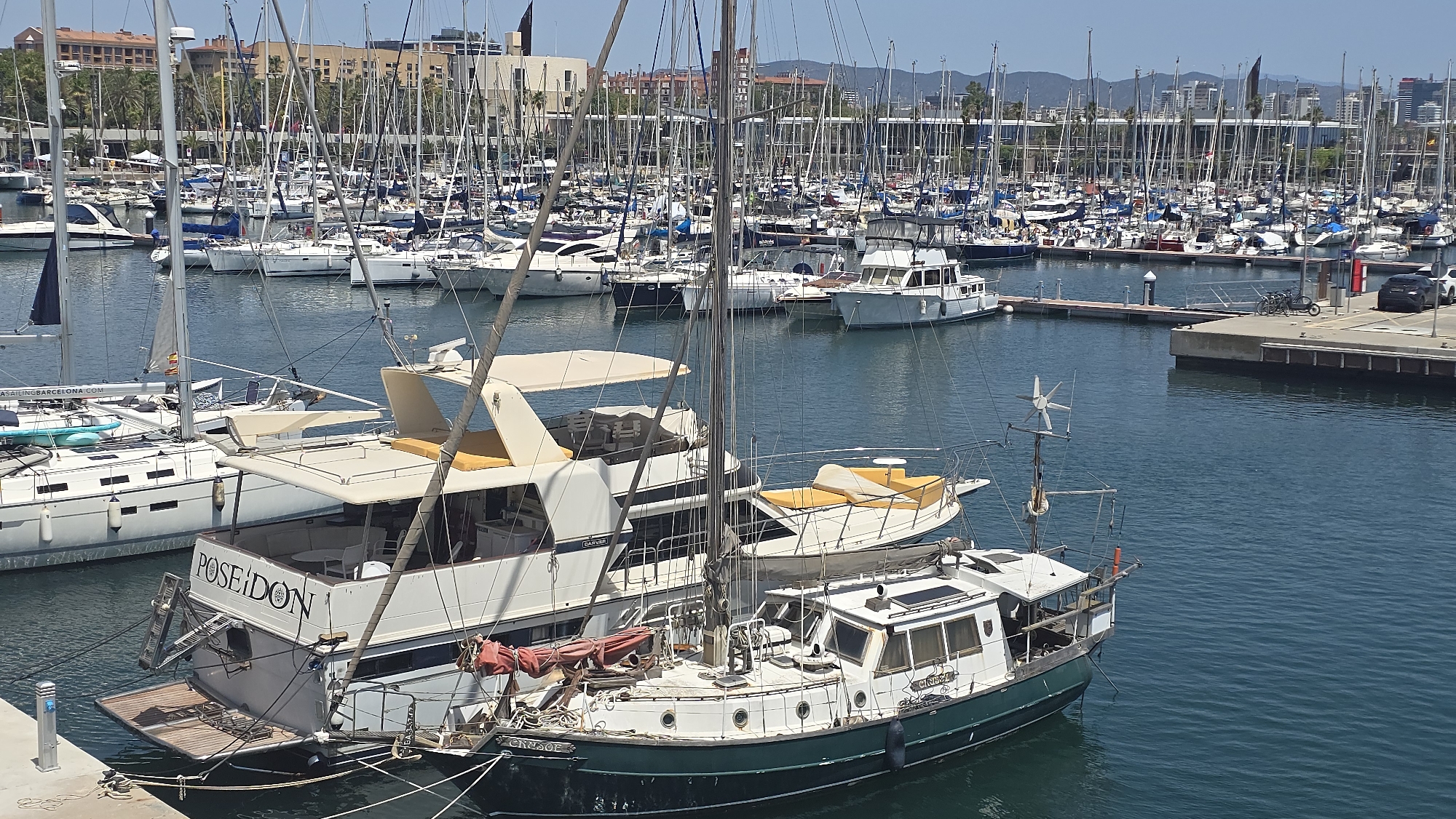 The image captures a sunny day at a marina in Barcelona, Spain, filled with numerous sailboats and motorboats docked in the blue water. The foreground features a large white yacht with "POSEIDON" emblazoned on its stern. Next to it, a smaller, older green and white boat with "ENSOF" visible on its side is also moored. In the background, a dense collection of sailboats with tall masts stretches towards the city skyline, where modern buildings and palm trees are visible. The sky is clear and bright, suggesting pleasant weather. The scene is a typical day at a busy harbor, with boats resting in the water under the daytime sun. There are no people readily visible in the frame. The presence of the marina and the city architecture strongly points to Barcelona as the location.