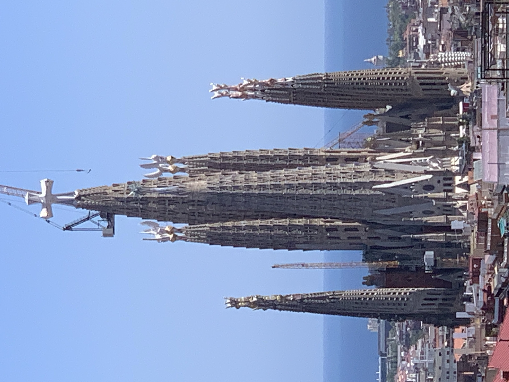 The image shows a distant, angled view of the Sagrada Familia basilica in Barcelona, Spain. The basilica is a large, unfinished stone structure with multiple spires, intricate architectural details, and scaffolding. A construction crane is visible near the top of one of the spires, indicating ongoing work. The sky is clear and blue, and the Mediterranean Sea is visible in the background, beyond the city's rooftops. The overall scene suggests a sunny day in an urban environment.