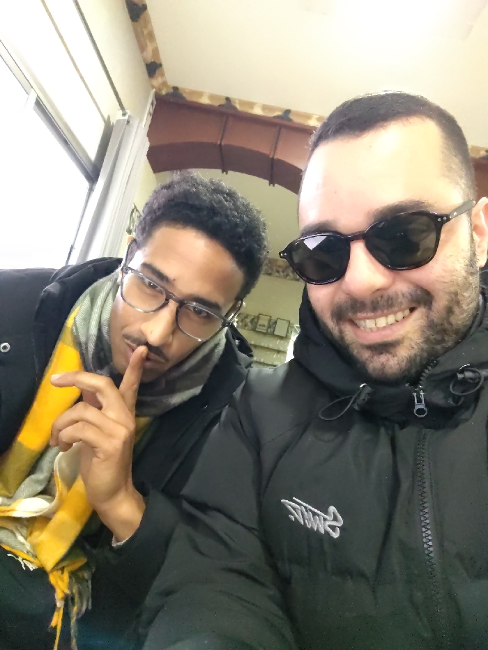 Two men pose for a selfie indoors. The man on the left wears glasses and a yellow and grey plaid scarf, holding a finger to his lips in a gesture for silence. The man on the right wears sunglasses and smiles, showing his teeth. Both are wearing black jackets. The background reveals a window on the left and an arched doorway.