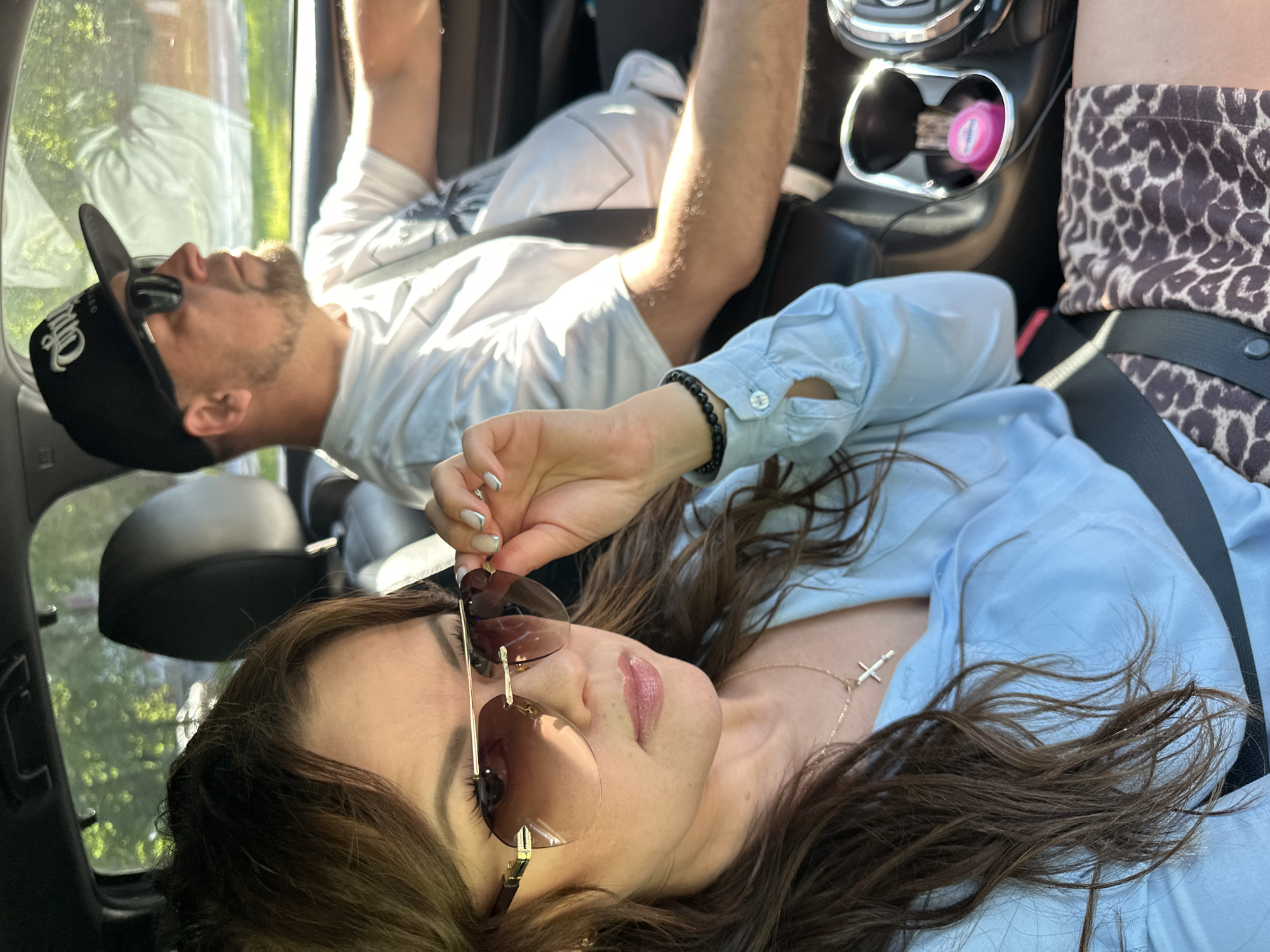 A car interior view shows two people, a male and a female, both appearing to be passengers. The female is in the foreground wearing sunglasses and a light blue shirt, secured with a seatbelt. She is holding her sunglasses near her face. A gold necklace is visible. The male is lying down in the background wearing a baseball cap, sunglasses, and a white shirt. A seatbelt is across his chest. The vehicle's console is visible, containing a pink container in a cupholder. The pattern on the other female’s leopard print pants is visible. The photo was taken in Chaikovsky, Russia.