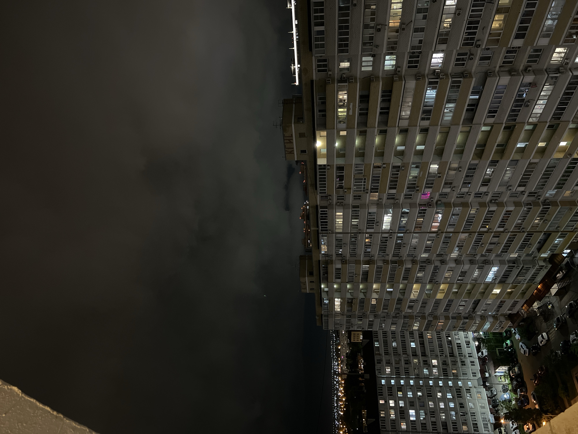 The image shows a high-angle view at night, possibly taken from a balcony, depicting a residential area in what is presumed to be the city of Krasnodar, Russia. On the right side of the image, several high-rise apartment buildings with many lit windows dominate the scene, indicating occupancy. The buildings are made of light-colored materials with external air conditioning units on many of the windows. Cars are parked on the grounds around the buildings.
To the left of the buildings, the night sky is overcast and dark. A smaller building is adjacent to the high-rise at the top. Some streetlights and faint city lights are visible in the distance between the buildings and the dark sky.