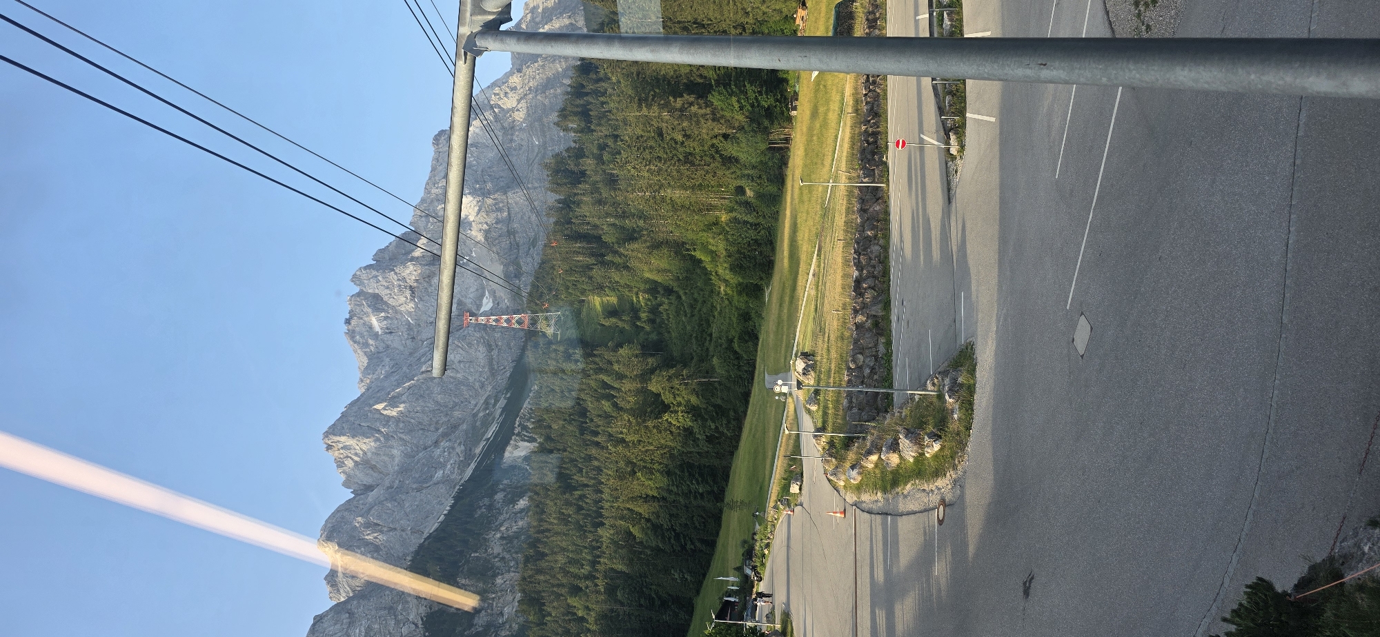 The image captures a scenic mountain landscape with a cable car system visible. In the foreground, a paved road curves through a parking lot, marked with white lines. A tall metal pole, part of the cable car infrastructure, stands prominently on the right. Beyond the road and parking area, a grassy slope rises, dotted with rocks and low walls. A dense forest of evergreen trees covers the lower slopes of the mountains. In the background, rugged, snow-capped mountains dominate the skyline, under a clear blue sky. A striking red and white pylon of the cable car system is positioned on the mountain slope, with several cable cars suspended from wires stretching across the frame. A bright, elongated light streak cuts across the upper left portion of the image, likely a reflection on the surface from which the photo was taken. The location is identified as Grainau, Germany, a Bavarian town known for its proximity to the Zugspitze, Germany's highest peak.