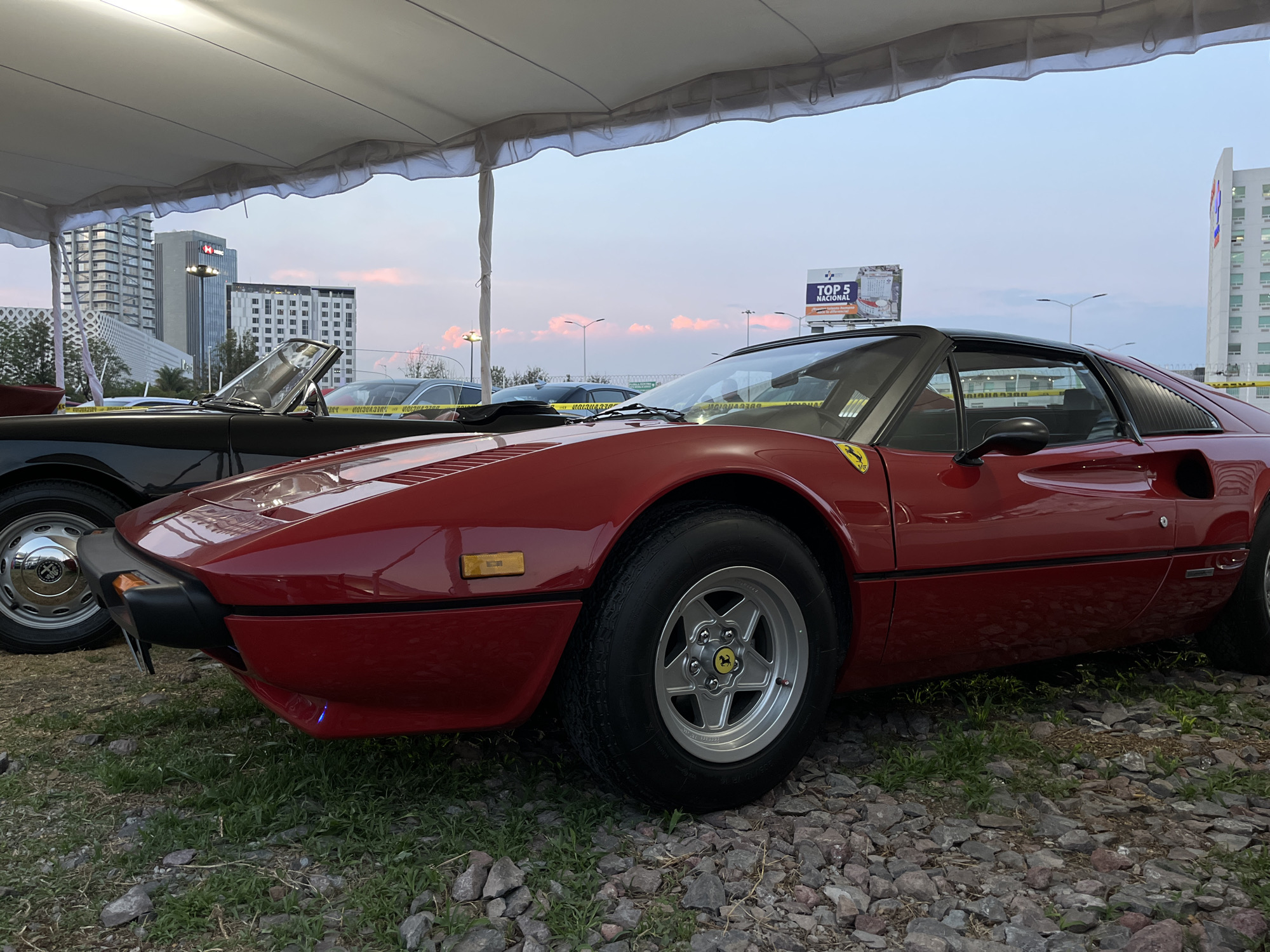 The image shows a red Ferrari 308 GTS parked on a gravel and grass surface, likely at an outdoor event. The car is the focal point, with its distinctive wedge shape and silver rims clearly visible. In the background, partially obscured, is a black classic car, possibly an Alfa Romeo Spider, also on display. Behind the cars, the urban skyline of Puebla, Mexico, is visible. Specifically, buildings such as the HSBC tower and other high-rise structures suggest the location is near the Angelopolis area. The sky is a muted evening hue with light pink clouds. Above the cars, a temporary white canopy tent is set up, providing shade. A billboard with the text "TOP 5 Nacional" is also visible in the background. The scene appears to be a vintage car show or exhibition.