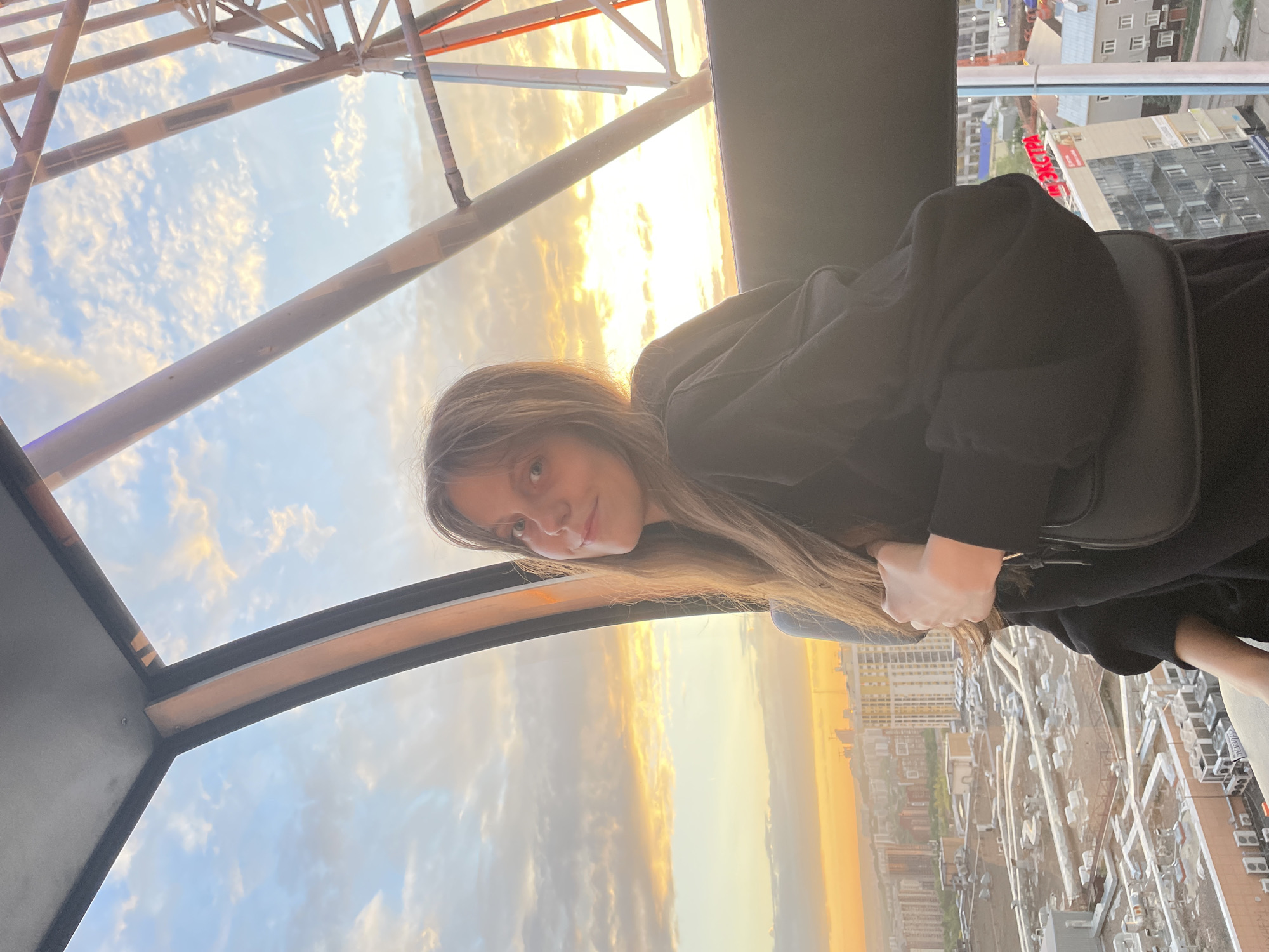 A young woman with long, light brown hair is seated inside a Ferris wheel cabin, looking towards the viewer with a slight smile. She is wearing a black hooded sweatshirt and carrying a black purse. The background shows a cityscape of Ufa, Russia, bathed in the warm light of a sunset. The sky is filled with scattered clouds, tinged with orange and yellow hues. The Ferris wheel structure is visible within the frame, along with the surrounding city buildings and infrastructure.