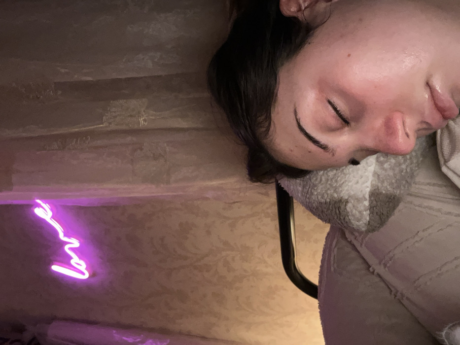 A person is sleeping on their side, head resting on a pillow, facing right. The room is dimly lit. A pink neon sign reading "love" is visible on the patterned wall. The individual has dark hair and closed eyes. The pillow is white and textured. A black frame is partially visible behind the pillow. The overall scene evokes a sense of peace and rest.