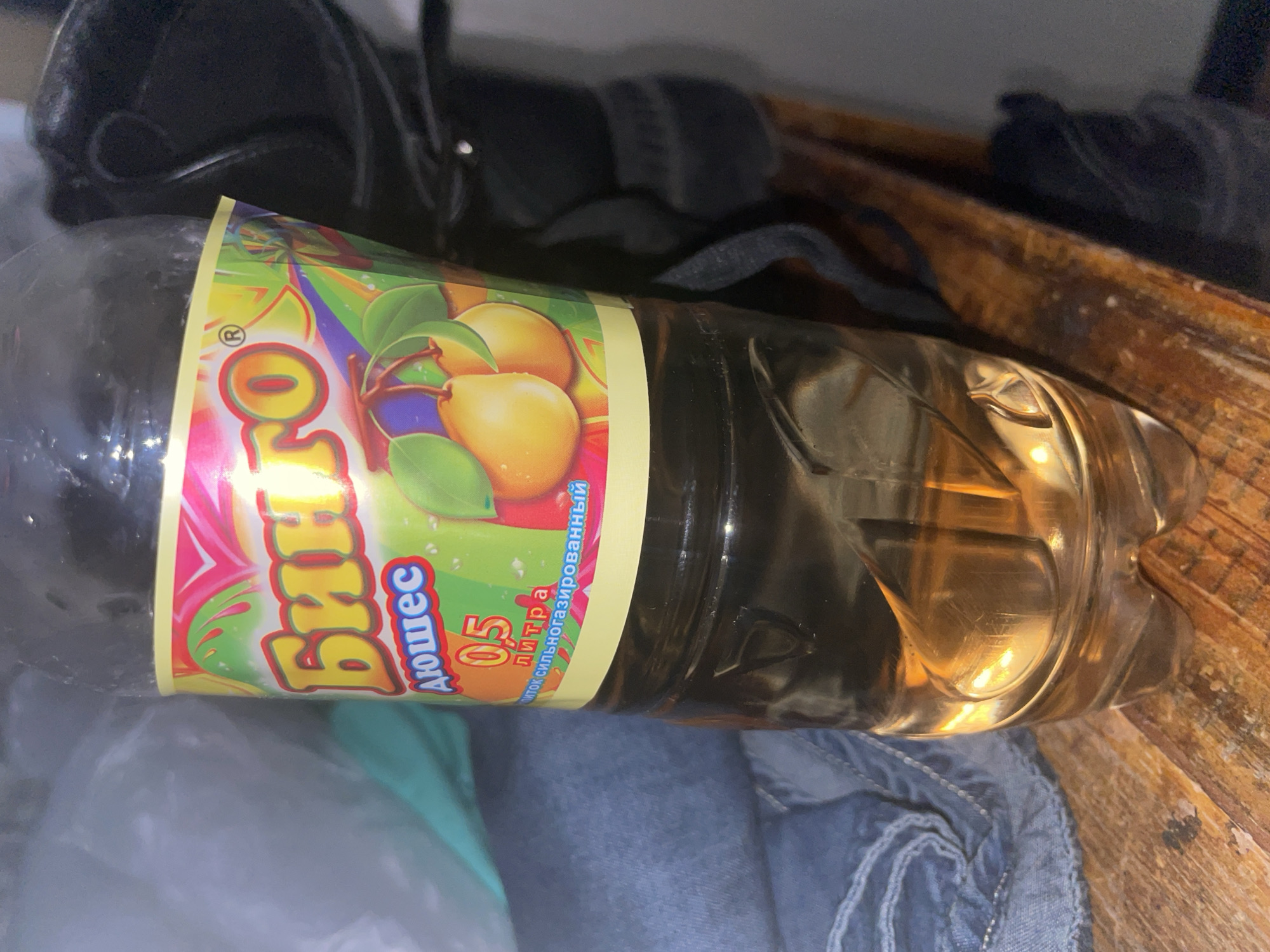 A close-up, eye-level shot shows a clear plastic bottle filled with a golden-amber liquid and a brightly colored label. The label features a cartoonish design with the word "Бинго" in large, yellow, outlined letters and the word "Дюшес" below it in smaller, blue, outlined letters. The number "0.5" is printed in red and orange, followed by "литра" in blue. Below this, in smaller blue text, it reads "питьсток сильногазированный." The bottle is positioned on a rustic wooden surface, with a stack of blue denim fabric to its left and dark, out-of-focus objects to its right. The lighting creates reflections on the surface of the liquid and the bottle. The background is dark and blurry, with hints of what appear to be clothing or bags.