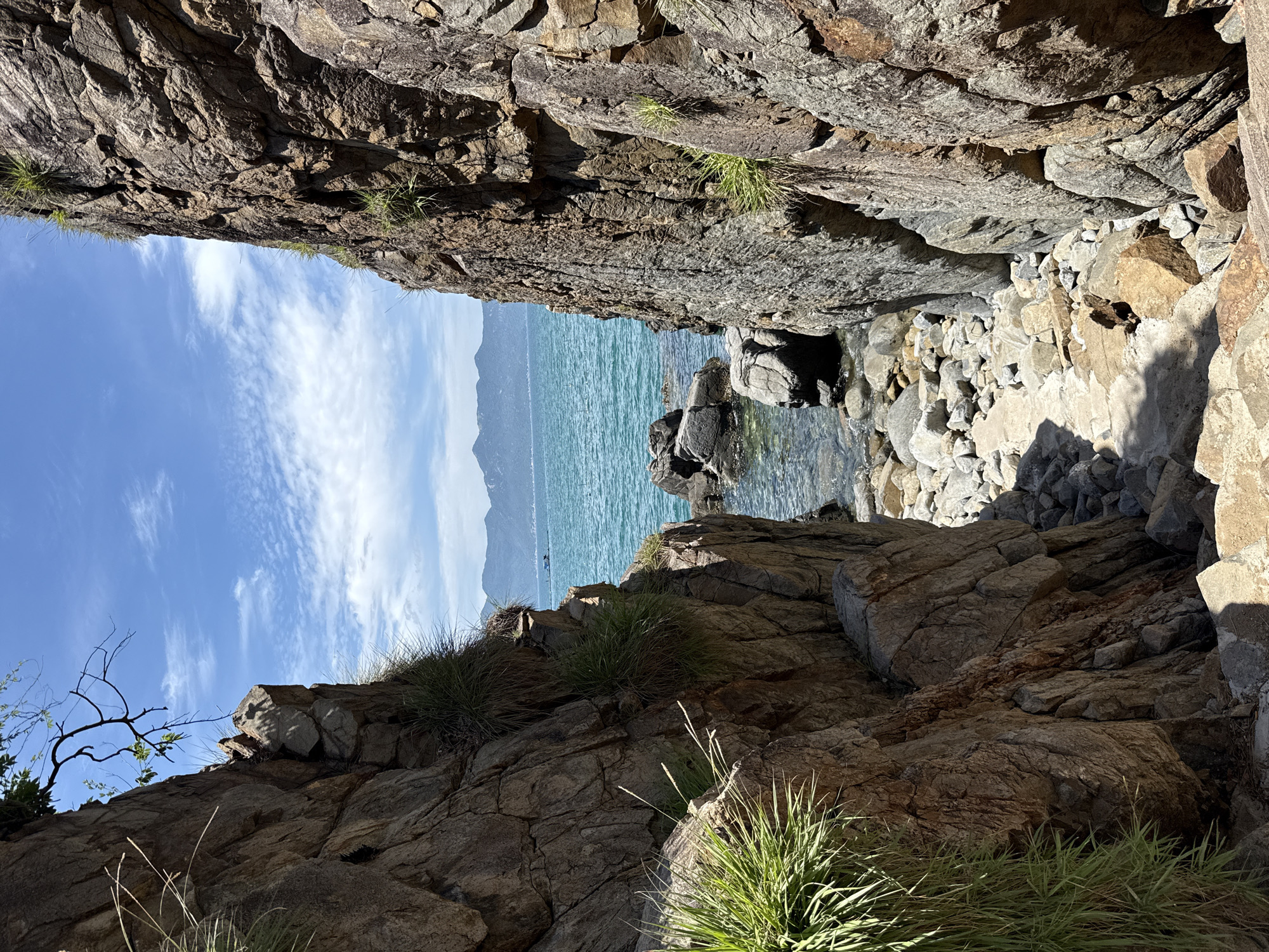 The image depicts a rugged coastal scene in Vĩnh Lương, Vietnam, with a narrow passage between two large, rocky cliffs leading towards the sea. The cliffs, composed of rough-hewn, brown and gray stone, are partially covered with sparse green grass and vegetation. The path leading down to the water is made of uneven rocks and pebbles. The turquoise sea is visible beyond the passage, with clear water revealing submerged rocks and a sandy bottom. In the distance, a hazy outline of mountains borders the horizon, under a bright blue sky dotted with scattered white clouds. A small boat can be seen on the water in the far distance. There are no people visible in the scene.
