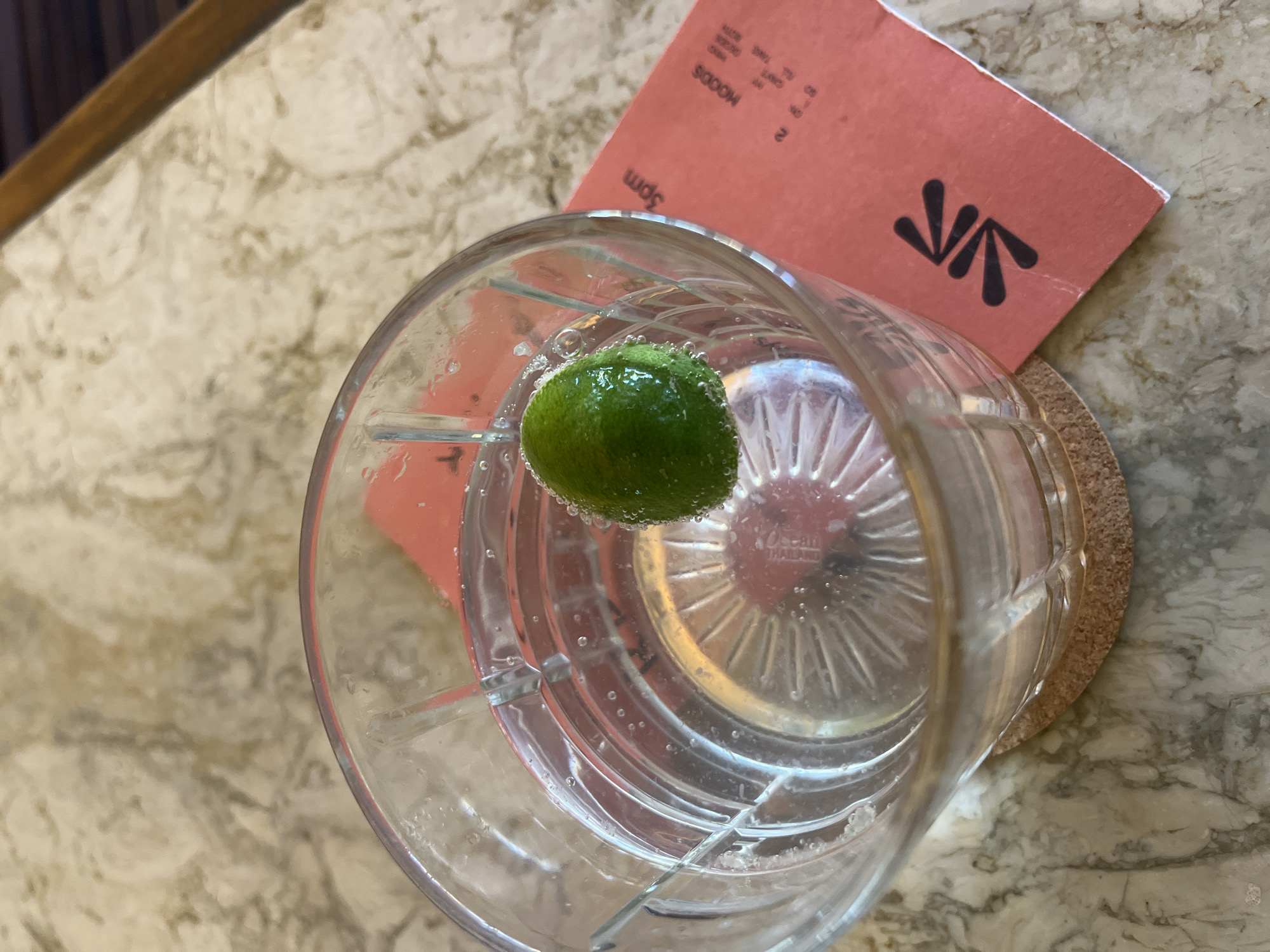 A close-up, high-angle shot reveals a clear, faceted glass filled with liquid and containing a green lime or lemon wedge. The glass sits on a coaster made of cork. Behind the glass and slightly to the upper right is a reddish-orange card with black printed text and a stylized logo. The glass and coaster rest on a marble-patterned surface. The restaurant is Moods, in North Kuta, Bali, Indonesia.