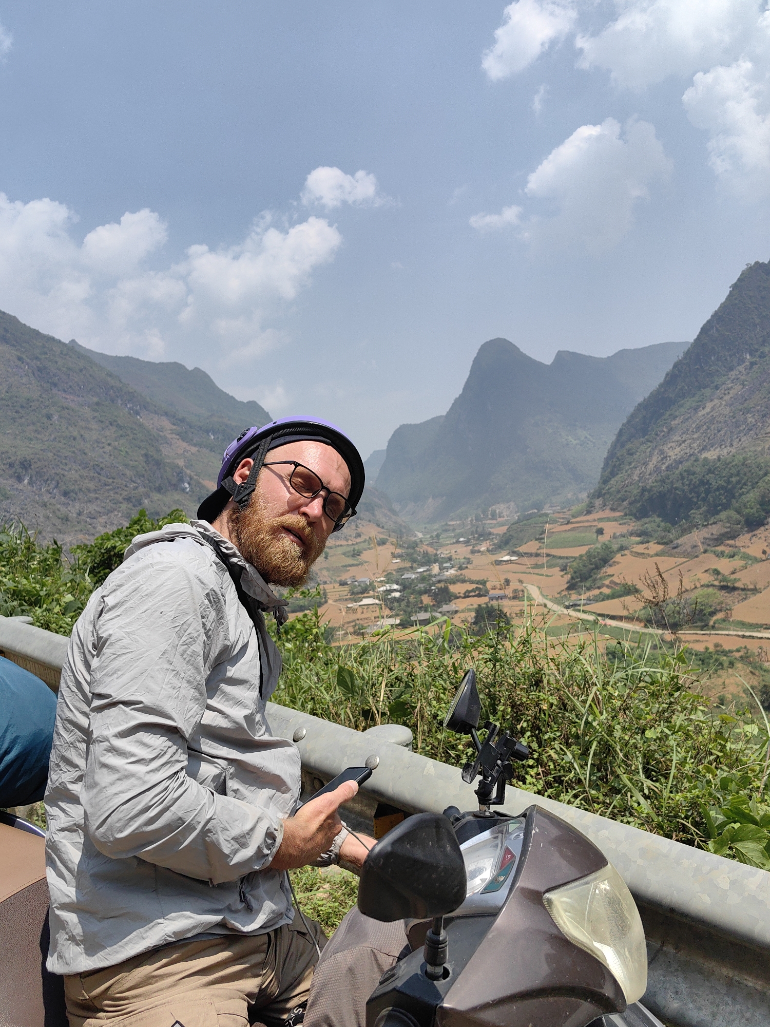 The image depicts a man on a motor scooter in Xã Xuân Trường, Vietnam. He is wearing a purple helmet, glasses, a grey jacket, and tan pants. He has a beard and is holding a smartphone. The scooter is brown and parked near a guardrail. Behind him is a scenic view of mountains and cultivated fields in the valley. The sky is blue with scattered clouds.