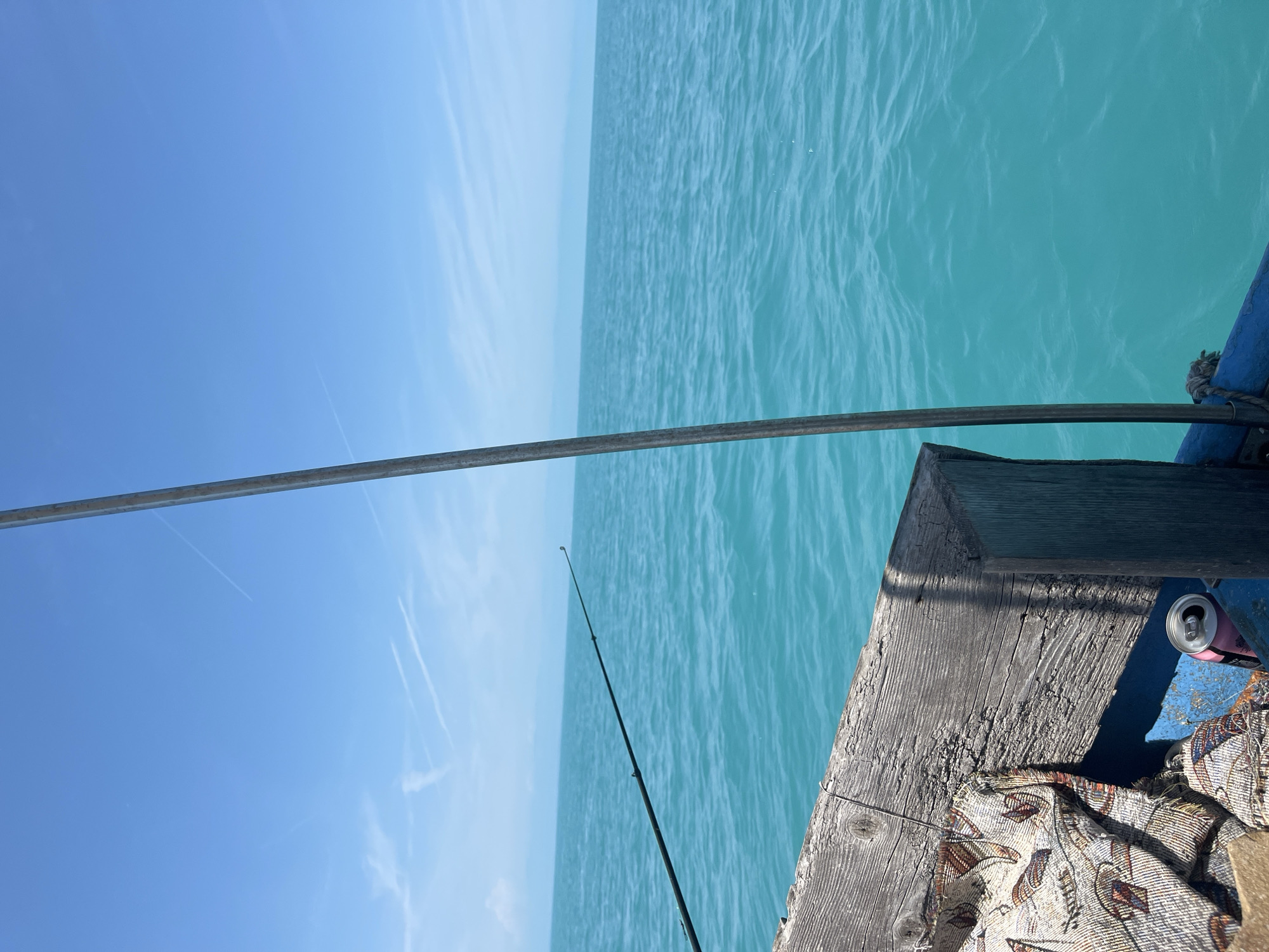 The image captures a detailed view from the port side of a weathered wooden boat, looking out onto a vast expanse of turquoise water under a clear blue sky, in the city of Unknown, Unknown.
The sky occupies the upper half of the frame, a vibrant light blue with several wispy white contrails, indicative of high-altitude aircraft. One prominent contrail stretches horizontally across the mid-upper right, while others are more diffuse or appear as fragmented streaks in the upper left. The horizon line, where the sky meets the water, is flat and distant, with a faint, hazy landmass visible in the far distance, particularly towards the right.
The water, which dominates the lower half of the frame, is a bright, clear turquoise with gentle ripples, suggesting calm conditions. No large waves are present.
In the foreground, the boat's structure is visible. A roughly textured, weathered wooden railing, bleached gray by exposure, runs horizontally across the bottom left quarter of the image. The wood shows signs of age, with some splintering and cracks. A piece of patterned fabric, possibly a blanket or tapestry with beige, orange, and blue hues in an abstract or leaf-like design, is draped over this wooden railing and into the boat's interior. Tucked beside the fabric in a darker blue-painted section of the boat's hull is an open, light pink aluminum beverage can.
A dark-colored fishing rod extends from the lower-left edge of the frame, angling upwards and outwards over the water towards the center-right. The tip of the rod is visible against the water and distant horizon. A thin, vertical metal pole, possibly a boat support or antenna, runs from the bottom edge of the frame to approximately two-thirds up the right side. A small, dark rope is visible tied to the boat's blue-painted hull section at the base of this pole on the right.
There are no people visible in the image. The scene suggests a tranquil moment of leisure and fishing on the water.