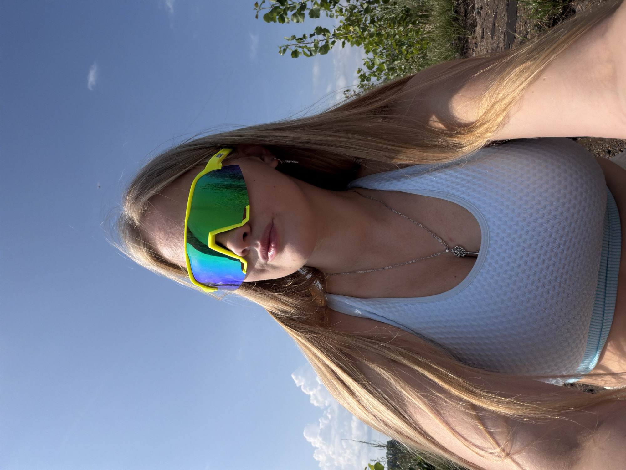 A young woman with long blonde hair, wearing a light blue sports bra and large yellow-framed sunglasses with green reflective lenses, is captured in a selfie. She is outdoors, with a clear blue sky and some scattered clouds above. To her right, there are green trees and vegetation. The ground appears to be a dirt or gravel path. A silver necklace with a key pendant is visible around her neck. The image was taken in Chaikovsky, Russia.