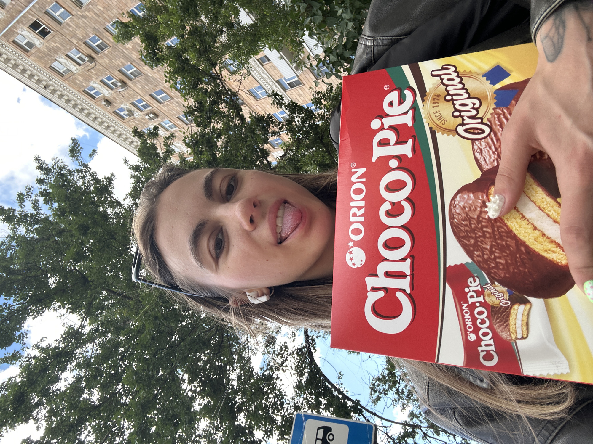 A woman is taking a selfie outdoors, holding a red box of Orion Choco-Pie. She is sticking her tongue out playfully at the camera, with her right hand holding the box and her left hand out of frame. Her left ear is adorned with a white earbud. Behind her, a tall, brick building with many windows looms, partially obscured by lush green trees. The sky is visible through the branches, with scattered clouds. A blue sign with a white bus symbol is visible at the bottom of the frame. The overall impression is a casual, spontaneous moment captured on a bright, possibly sunny day in an urban environment. The text "ORION Choco-Pie Original" is prominently displayed on the box.