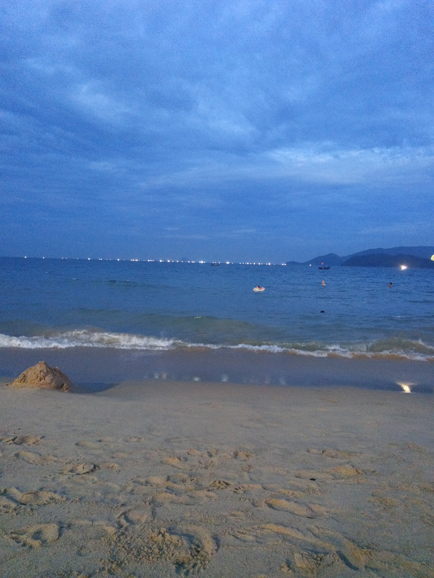 This media file presents a serene beach scene at dusk or early night in Nha Trang Ward, Vietnam. The sky above is a deep, cloudy blue, with muted light indicating the transition from day to night.
In the foreground, a sandy beach fills the lower half of the frame. A small, organic-shaped sand mound or sandcastle is visible on the left, close to the waterline. Numerous footprints and textures are imprinted across the damp sand, subtly illuminated.
The midground features the calm ocean with gentle white waves breaking softly onto the shore. Several individuals are visible enjoying the water. One person floats on a bright yellow inflatable ring, while at least two other figures are partially submerged, seemingly swimming or standing in the sea. Small boats and numerous distant lights punctuate the horizon across the water, likely from other vessels or a faraway coastline. One specific boat can be seen closer to the distant landmass.
In the background, a line of distant mountains is silhouetted against the dark sky on the right side of the horizon. The overall lighting is dim, emphasizing the artificial lights reflecting on the water. The scene conveys a peaceful atmosphere, depicting leisurely evening activities by the sea. There is no visible text in the image.