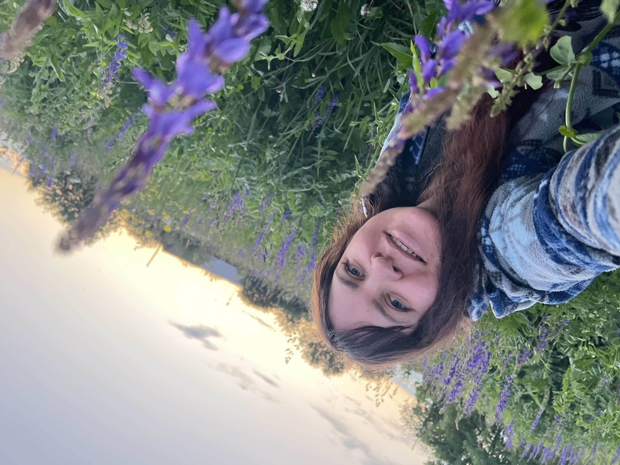 A woman with long, brown hair and bangs smiles at the camera in a field of purple flowers. She is wearing a blue and white patterned jacket. The background is a blur of green foliage, purple flowers, and a hazy sky with clouds. The image is taken from a low angle, looking up at the woman. The location is identified as Panteleevka, Russia.
