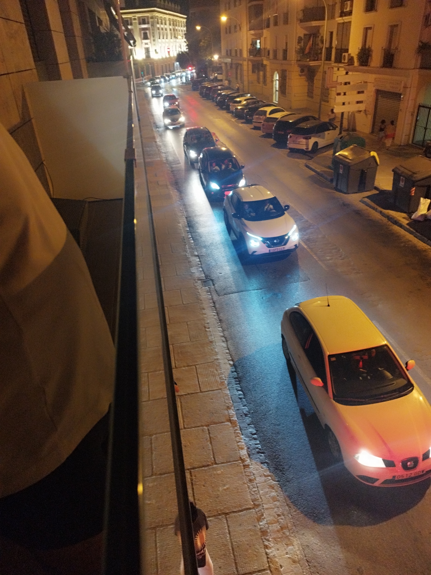 The image captures a nighttime street scene in Jerez, Spain, characterized by a row of parked and moving cars. The perspective is from an elevated viewpoint, looking down onto a street paved with light-colored bricks. A few buildings with illuminated windows line the right side of the street, suggesting a commercial or residential area.
In the foreground, a light-colored car, possibly a hatchback, is prominently featured. It has its headlights on, illuminating the street. Further down the street, a white SUV is seen with its headlights on as well, indicating it is either moving or stopped with its lights activated. Behind it, other cars are visible, some parked along the curb and others in motion. The street is wet, reflecting the lights from the vehicles and buildings, suggesting recent rain or a damp environment.
The presence of streetlights and illuminated building facades indicates an urban setting after dark. The overall impression is one of a typical city street at night, with traffic and urban infrastructure visible.