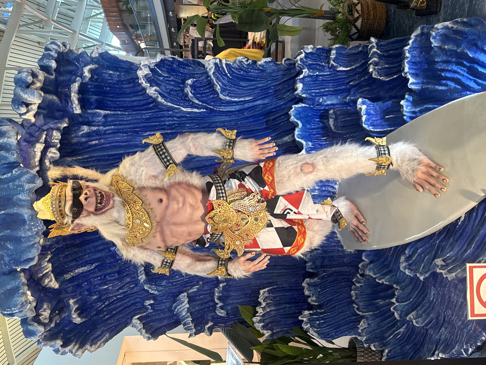 The image captures a statue of Hanuman riding a surfboard amidst a backdrop of stylized blue waves. The statue is adorned with Balinese attire, including a headdress, decorative chest piece, and wristbands. The statue's facial expression is exaggerated, with large teeth and a prominent nose. The setting appears to be indoors, potentially an airport or commercial space, based on the overhead structure. This statue is a popular tourist attraction, often found in Kuta, Indonesia.