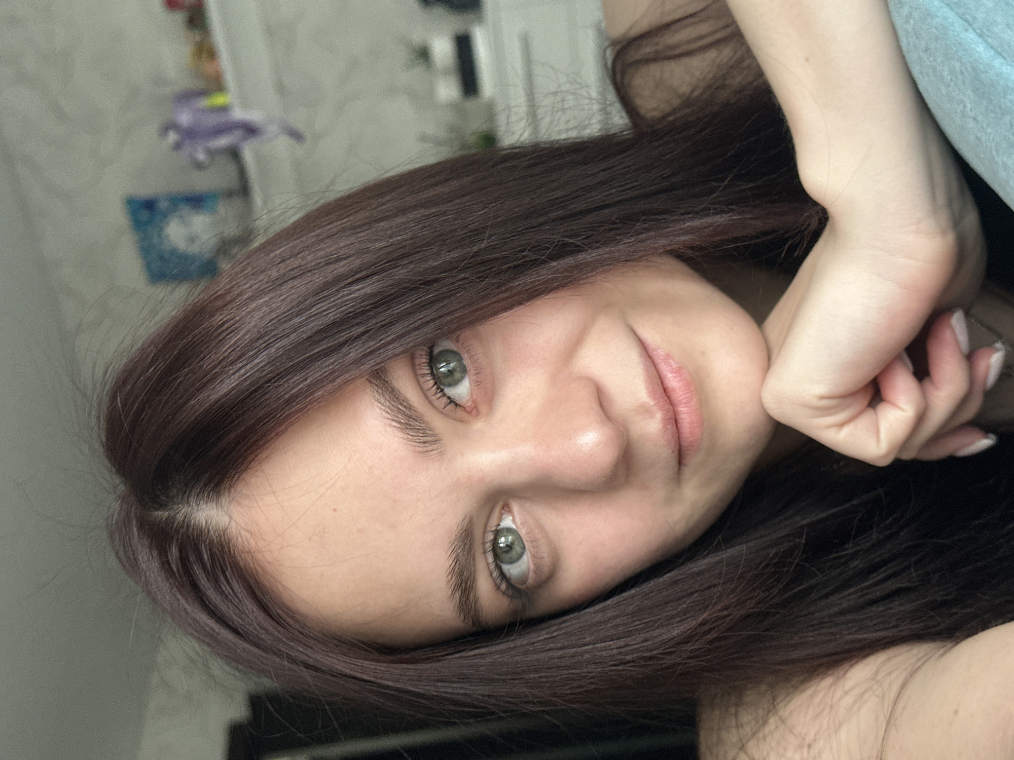 A young woman with long, dark brown hair and green eyes is captured in a close-up, diagonal selfie. Her face, framed by her hair, looks directly at the camera with a gentle, slight smile, conveying a calm expression. Her right hand is visible, resting with her chin supported, and her nails are painted with a light-colored polish that appears somewhat worn.
The background is softly blurred, indicating an indoor setting, likely a home. Faintly visible in the upper left corner are indistinct shapes, including a small, blurry light purple object (possibly a toy or decoration) and a blue rectangular object below it, which could be a framed picture or a small sign. Further to the right, parts of what might be furniture or a door frame are obscured. Light blue or grey fabric, possibly clothing or a blanket, is visible in the upper right. The lighting suggests it is daytime, with soft, diffused light illuminating her face. There is no visible text in the image. The image itself is rotated approximately 90 degrees clockwise.
