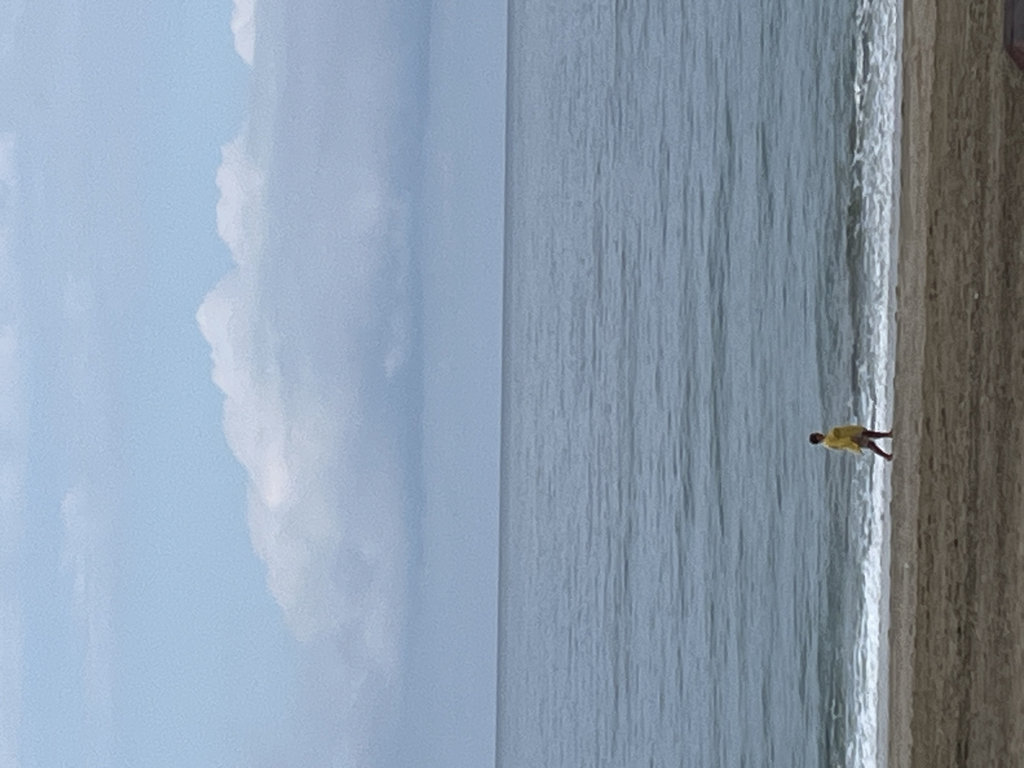 The image presents a beach scene in Alanya, Turkey. A figure, presumably a person, stands on the sand near the water's edge. The ocean spans a large portion of the image, its surface reflecting light. The sky is visible on the left, filled with clouds.