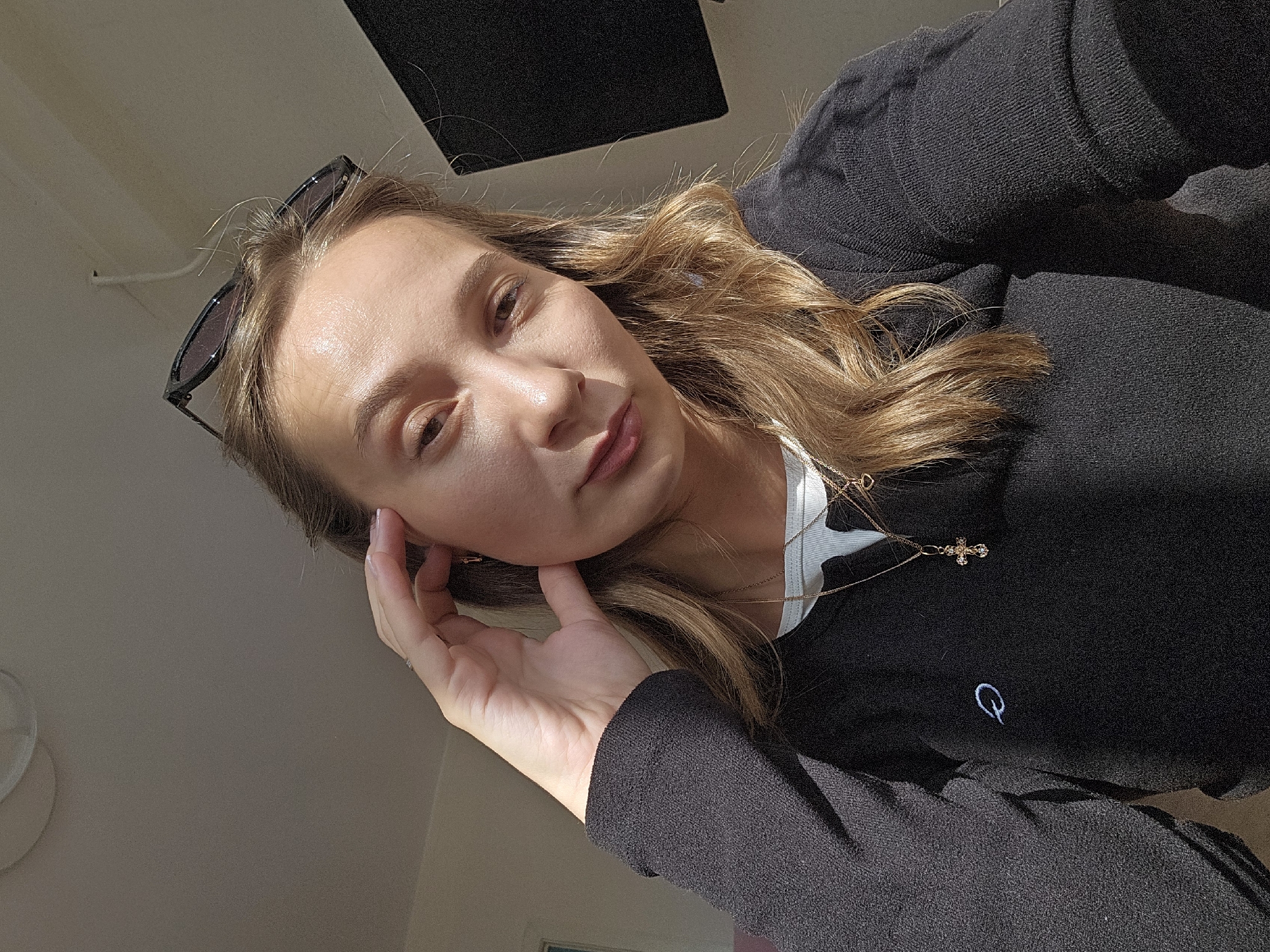A selfie shows a woman indoors, possibly in Chaikovsky, Russia, based on contextual knowledge. She is fair-skinned with brown hair, wearing a black sweater and a gold necklace with a cross pendant. Sunglasses are perched on her head. Her left hand is near her face, and a small "Q" logo is visible on the sweater. Natural light illuminates the scene, creating shadows. A dark, rectangular object is visible above her.