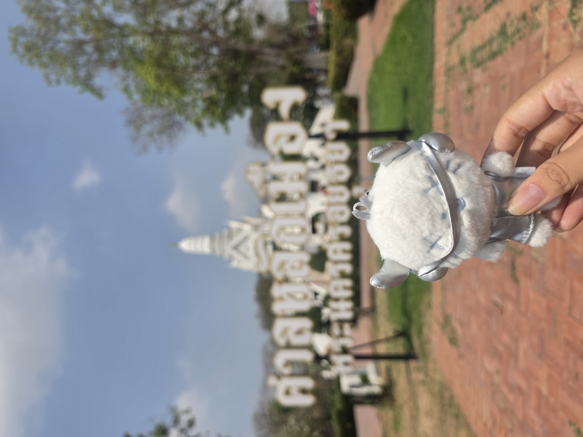 A hand holds a small, white stuffed animal against a background that features a large sign with Thai script and a white pagoda-like structure. The location is the Historic City of Ayutthaya, Thailand. The background also includes trees, grass, a clear blue sky, and brick walkways.