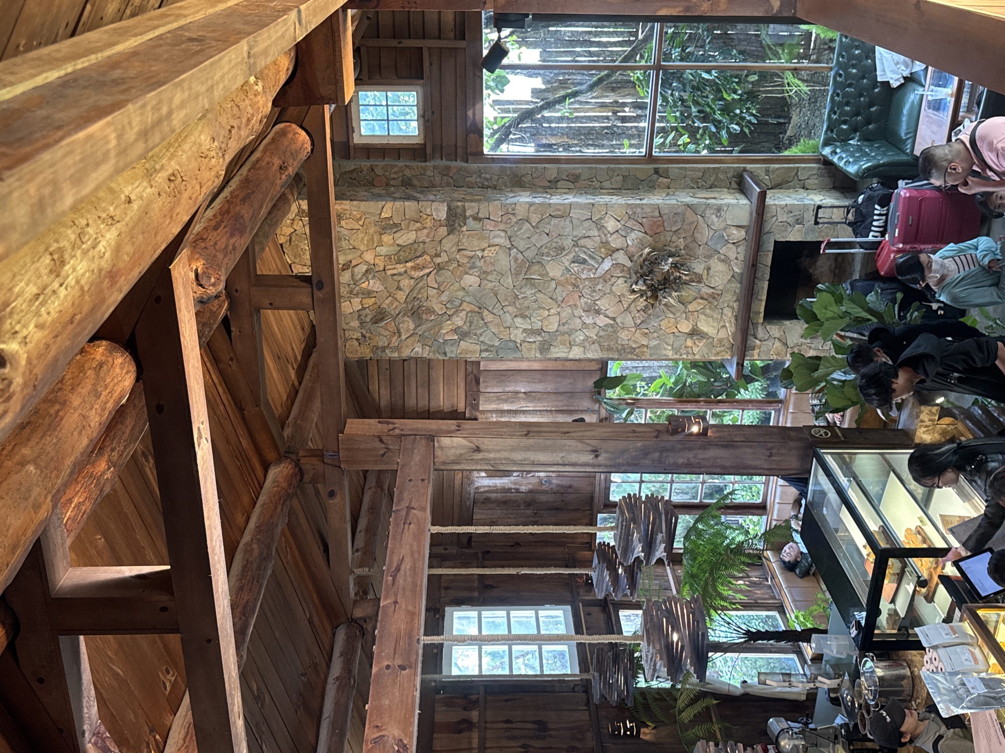 The image depicts the interior of a rustic, wooden coffee shop, likely located in Da Lat, Vietnam, given the prevalent style of architecture and ambiance. The space features high, exposed wooden beams and a stone fireplace. Several large windows offer views of lush greenery outside.
In the foreground, a counter area is visible with various coffee-making equipment and a display case filled with pastries. Several people are gathered around the counter, appearing to be customers or staff. One person, wearing a black baseball cap and an apron, is standing behind the counter. Another individual, wearing glasses, is interacting with a person in front of the counter.
Further into the shop, a green leather sofa is placed near the fireplace, with luggage, including pink suitcases and bags, next to it. A person is seen relaxing on a raised platform or banquette by a window. The overall atmosphere is relaxed and cozy, suggesting a popular spot for patrons to unwind.