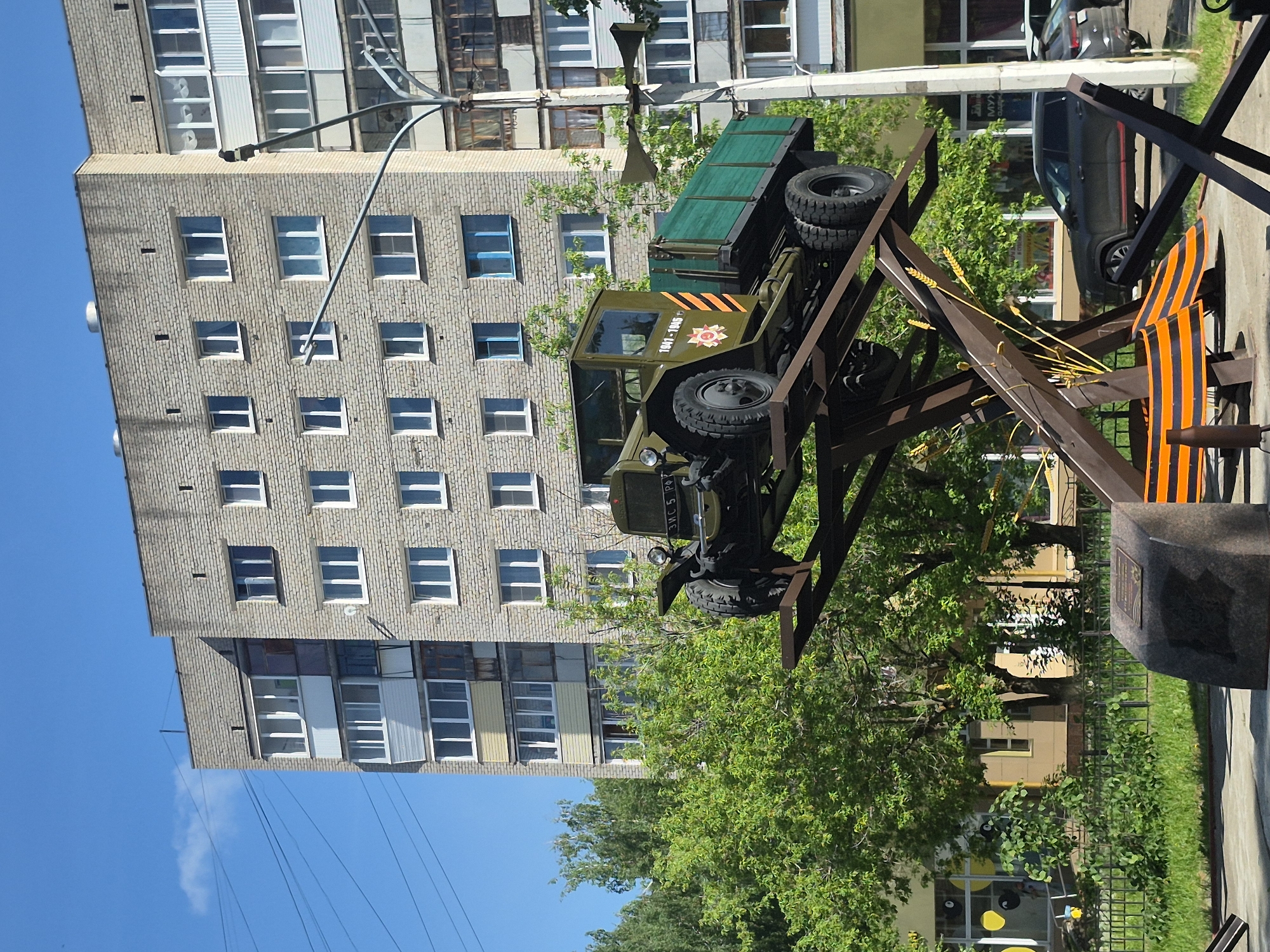 A monument featuring a military vehicle is erected in an urban area, likely in the city of Unknown, Unknown. The green military truck is mounted on a dark brown metal structure, which also features wheat stalks and a black and orange striped ribbon. A stone plaque sits alongside the structure. The background contains a multi-story building with a repetitive window pattern and green foliage. A blue sky is visible at the top left corner of the frame.