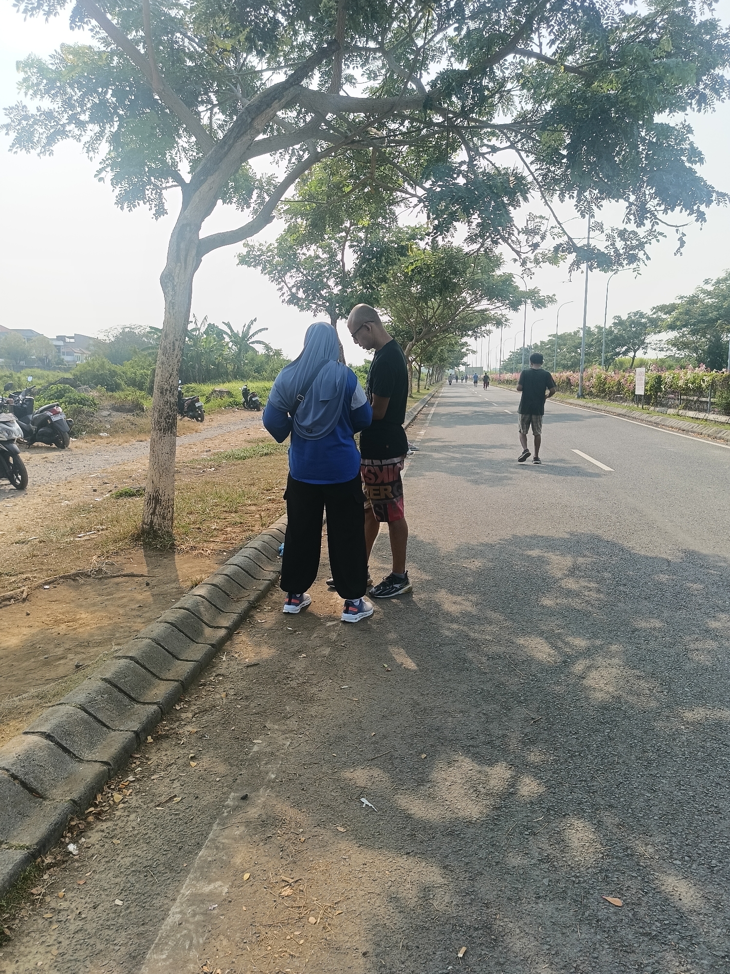 In Punggul, Indonesia, a dry, dusty roadside scene unfolds under a bright, hazy sky, shaded by the dense canopy of a large tree. On the paved road, a man in a black t-shirt and patterned shorts jogs away from the viewer, his back to the camera. Further down the road, several other figures are visible, seemingly engaged in outdoor activity. Beside the road, two individuals stand facing each other. One, wearing a blue hijab and a blue top with dark pants, appears to be looking at a phone. Beside them, a man in a black t-shirt and patterned shorts also looks down, possibly at a phone. Their interaction is brief and focused. To the left of the road, several motorcycles are parked on a dirt and grass area, suggesting a possible mode of transport for those in the vicinity. The environment appears to be on the outskirts of a populated area, with hints of residential buildings and vegetation in the background, characteristic of a suburban or semi-rural setting in Punggul. The overall atmosphere is casual and active, hinting at people making use of the outdoor space.