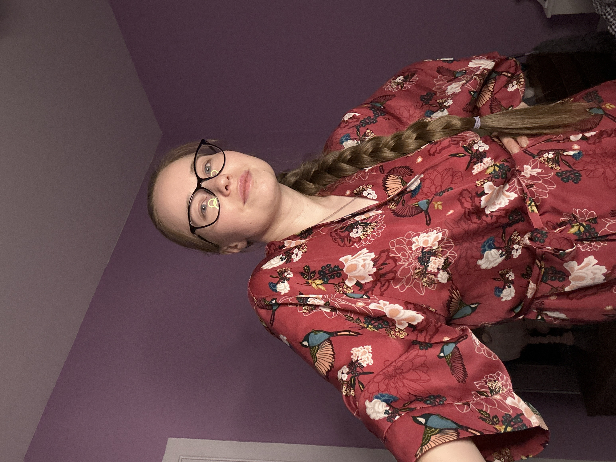 A woman with a blonde braid and wearing black framed glasses is shown from a low angle, in a photograph taken indoors. She is dressed in a red floral patterned robe. The background consists of a purple wall, a bed, and a white door frame near the bottom of the frame. The location is not identifiable as Saint Petersburg, Russia, based solely on the provided image.