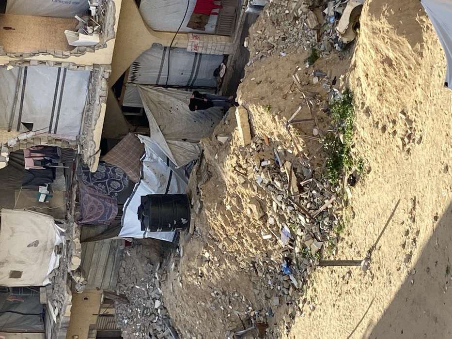 The image depicts a devastated urban area in Khan Yunis, Palestinian Territory, characterized by extensive structural damage and makeshift living arrangements. The foreground and mid-ground consist of large, irregular mounds of light brown sand heavily intermixed with construction debris, including concrete fragments, bricks, and twisted metal. A black, cylindrical water tank is centrally positioned on one such sand pile, partially obscured by a white tarp draped over an adjacent makeshift structure.
In the background, a multi-story concrete building shows significant damage, with large sections of its facade missing, exposing internal components and rebar. Numerous white and patterned tarpaulins, blankets, and fabrics are draped over the damaged sections of the building and utilized as temporary walls and coverings for makeshift shelters at ground level. A blue UNICEF logo is visible on a white tarp covering a part of the building on the upper right. Various articles of clothing, including a red garment, are hung to dry from the building's damaged structure.
One individual is present in the mid-ground to the right, dressed in dark clothing, bent over near a makeshift entrance formed by a white tarp. The person's face is obscured, and their specific action is indeterminate. Small patches of green vegetation grow sporadically amidst the sand and rubble. A dark, slender pole stands vertically in the lower-left foreground, casting a shadow on the sandy ground.