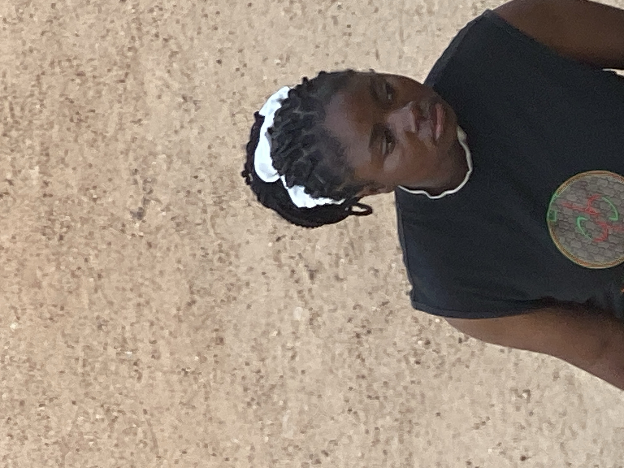 A close-up, medium shot captures a young Black woman from the chest up, positioned on the right side of the frame. She is looking down and to the right, her face slightly turned away from the camera. Her braided dark hair is pulled back and adorned with a white scrunchie. She wears a sleeveless black t-shirt with a white trim around the neckline. A circular emblem featuring red and green motifs is visible on the front of her shirt. The background is a textured, light brown surface, possibly a wall or ground. The image appears to have been taken outdoors in Jalingo, Nigeria.