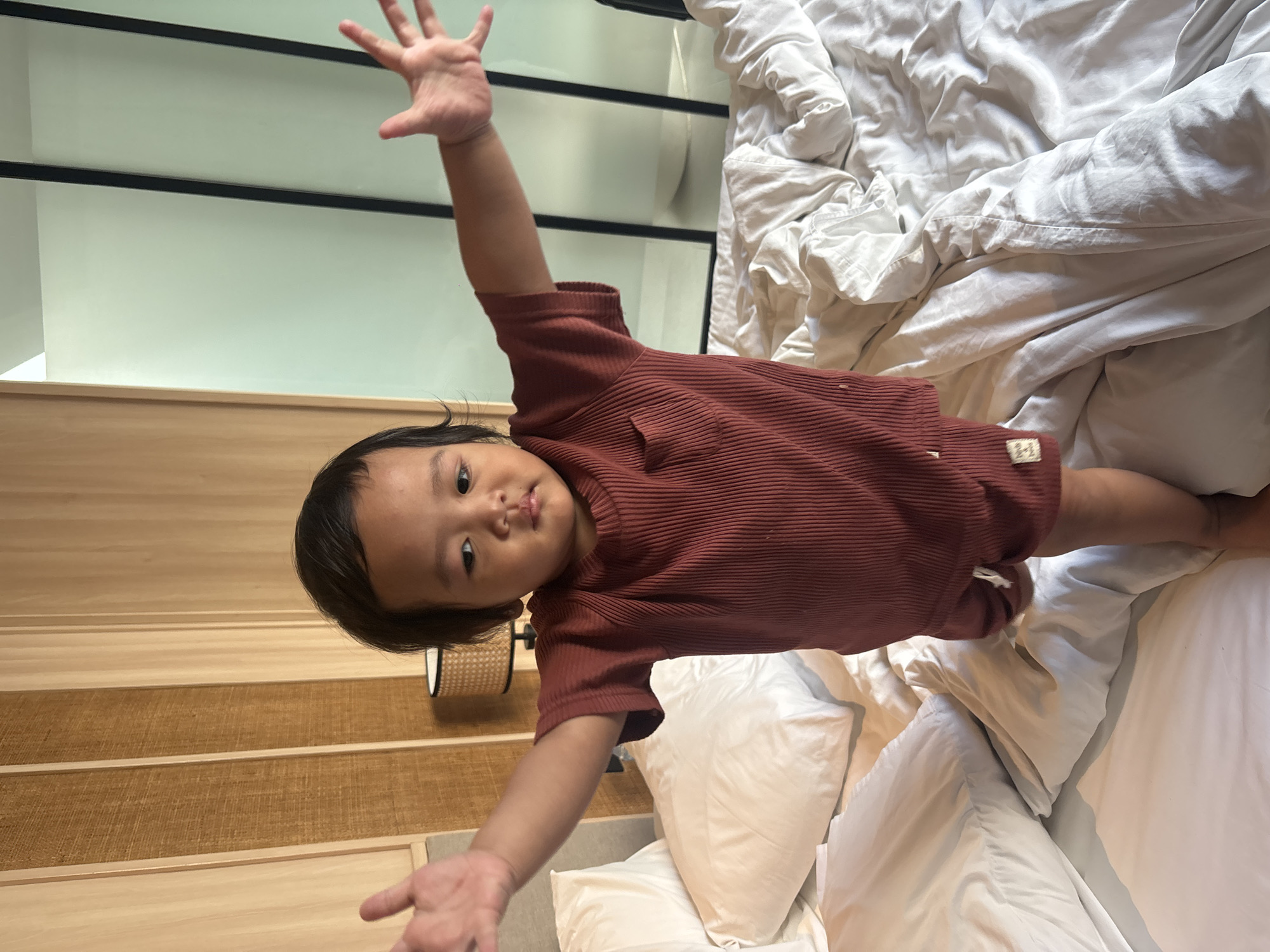 In North Kuta, Indonesia, a young child stands on a bed with their arms outstretched. The child is wearing a reddish-brown ribbed t-shirt and shorts. The bed is covered in rumpled white sheets. In the background, there is a wooden headboard and a wall with vertical panels. A bedside lamp with a woven shade is visible to the left. The overall environment suggests a hotel room or a residential bedroom.