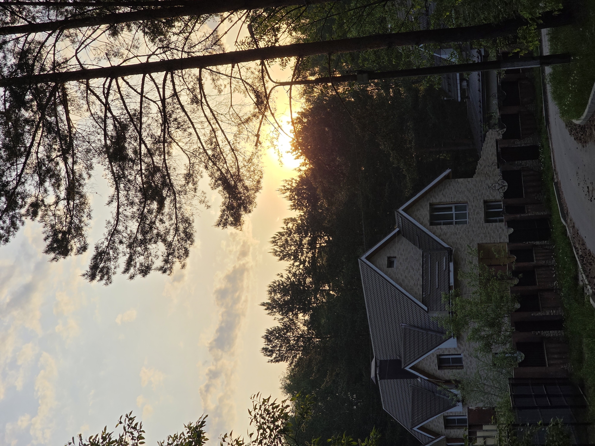 The image shows a sunset in what appears to be a suburban or rural area. The sun is low on the horizon, partially obscured by trees. The sky is a pale yellow color. In the foreground, the branches of a tree are silhouetted against the sky. A house is visible in the middle ground, constructed of light-colored brick with a gray roof. A decorative metal fence runs along the side of the house. A paved road runs along the right side of the image. The location is likely Chaikovsky, Russia, based on the user's prompt. No people are visible in the image.