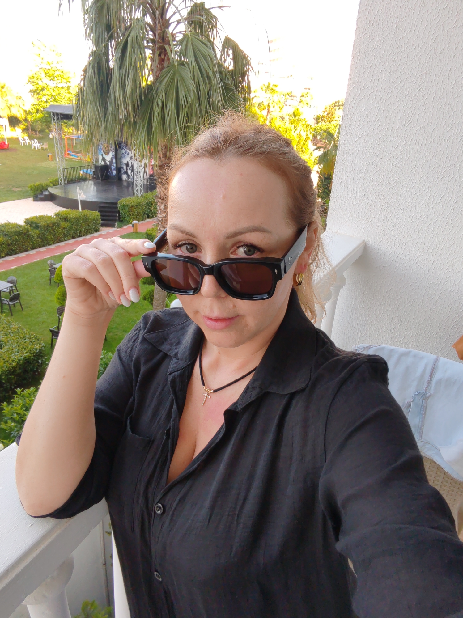 A woman stands on a balcony, looking into the camera while adjusting black sunglasses with her left hand. She is wearing a black button-down shirt and a delicate necklace with a small cross pendant. She has fair skin, light-colored hair pulled back, and gold hoop earrings. The background shows a resort-like area with green lawns, trees, and a small outdoor stage. Based on the resort-style environment and possible presence of Mediterranean flora, there is a strong possibility that this photo was taken in Alanya, Turkey.