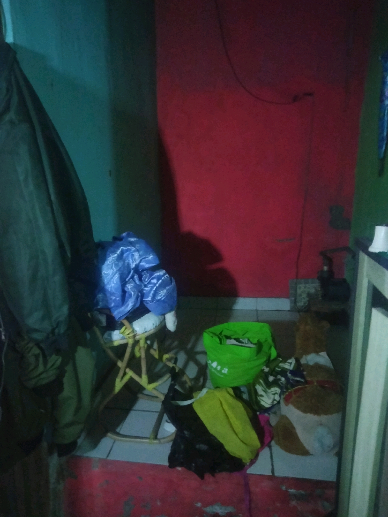 The image depicts a dimly lit, cluttered interior space. On the left, a dark green garment, possibly a jacket or coat, hangs from a hook or railing, obscuring the wall behind it. To its right, a small stool or chair is visible, topped with a pile of blue fabric, likely clothing, with a patterned design. Further to the right, a bright green fabric bag stands upright, partially filled. In front of the green bag, there are several other items of clothing or fabric, including a yellow and black bundle, and a pink cloth. To the right of these items, a round object with brown and white patterned fabric, resembling a cushion or a stuffed toy, is visible. In the background, the walls are painted red on the right and a teal-green on the left. The floor appears to be tiled, with a red baseboard running along the bottom of the walls. A small black pump or water filter device is visible near the red wall on the right. The lighting in the room is poor, casting shadows and contributing to a generally disheveled appearance. The scene appears to be indoors, possibly a storage area or a bedroom. The location is specified as Suka Sari, Indonesia.