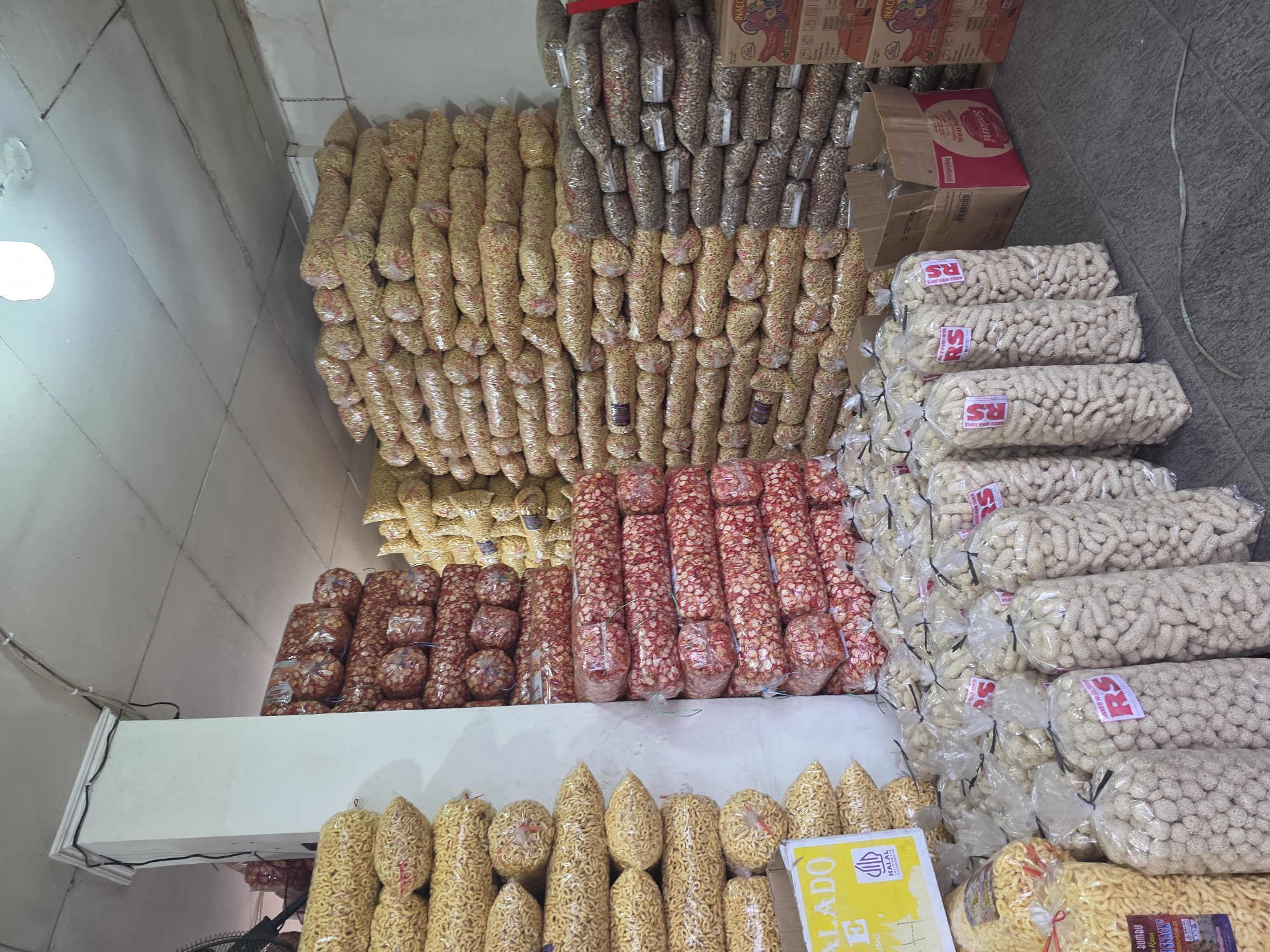 The image depicts a storage area, likely a warehouse or a market stall, filled with numerous large, clear plastic bags of snacks. The bags are stacked high, forming walls of packaged goods. The snacks appear to be of various types, with some being elongated and yellow, others round and red, and a third type small and off-white, possibly coated in a light-colored powder or glaze. Many bags have red tags with the letters "RS" printed on them, and some yellow bags have informational labels, one of which includes the "HALAL INDONESIA" certification mark. In the foreground, several stacks of the off-white snacks are prominently displayed. In the background, more stacks of yellow and red snacks are visible, along with some cardboard boxes. The environment is dimly lit, with a single fluorescent light fixture visible on the ceiling. The flooring appears to be tiled. The overall impression is of a bulk snack distribution or retail point within a commercial area. Based on the "HALAL INDONESIA" certification and the context of snack distribution, this scene is consistent with a location in Mekarsari, Indonesia. There are no people visible in the image.