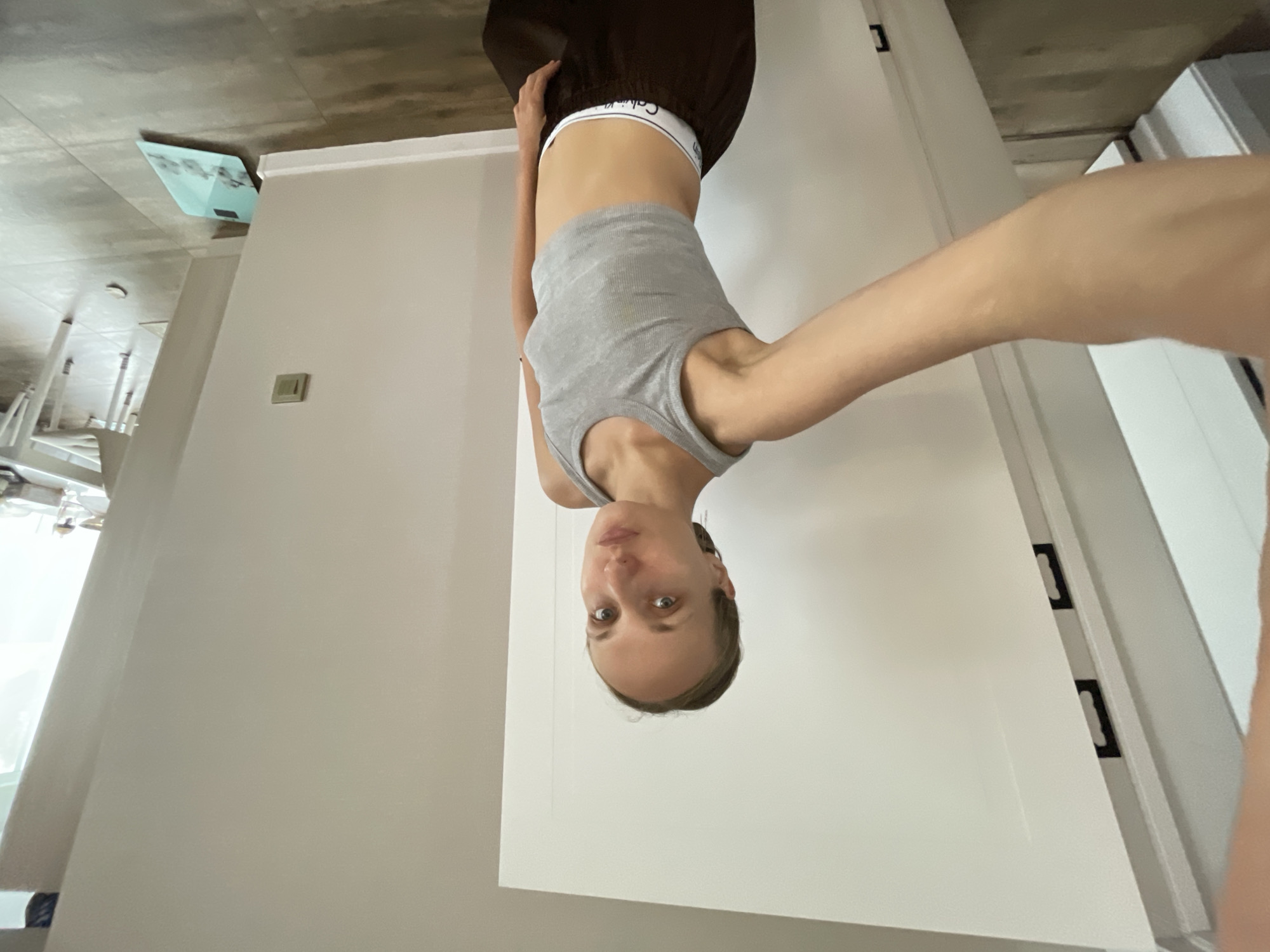 A young woman with blonde hair pulled back is taking a selfie in what appears to be a home environment. She is wearing a gray tank top and brown pants. The background includes a white door and a wall with a light switch. In the distance, a table and chairs can be seen, suggesting a dining area. There is also a blue dustpan on the floor. The overall impression is casual and domestic. It is not possible to determine the location as the city of Подшивалово, Russia.