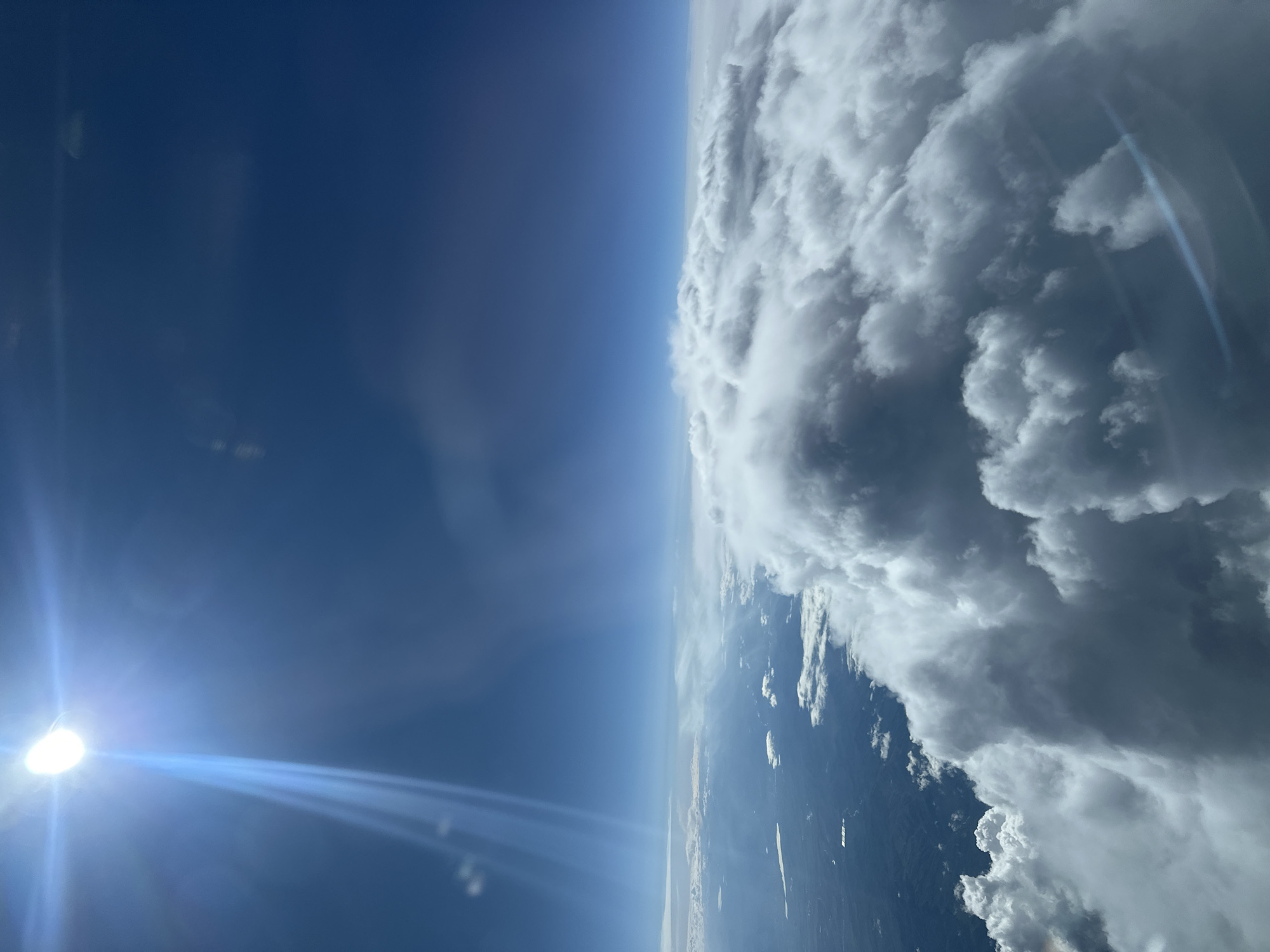 The image shows a view from an airplane window. The left side of the frame is a clear, blue sky with the sun visible. A bright light flare emanates from the sun, stretching across the sky. The right side of the frame shows a dense formation of clouds with visible land below. There is no evidence to confirm it is the city of Khimki, Russia.