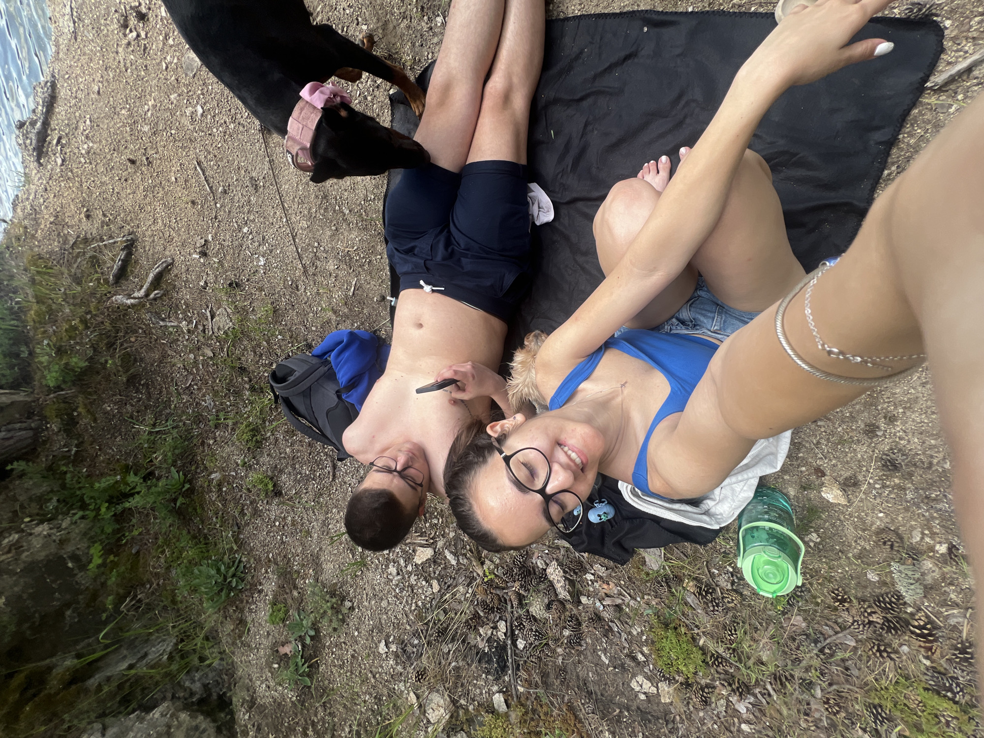 This image depicts a high-angle selfie taken outdoors in a natural setting, identified contextually as Alino, Bulgaria. Two individuals and a dog are positioned on a gravelly and dirt ground with sparse vegetation and numerous pine cones. A body of water is visible in the upper right background, with a rocky shoreline and visible ripples.
In the foreground, a female with light skin tone, dark hair, and eyeglasses is smiling and looking towards the camera, holding the device with her right arm. She is wearing a blue swimsuit top, light blue denim shorts, and has white nail polish on her toenails. A silver chain bracelet adorns her right wrist. A large, translucent green water bottle with a light green lid rests on the ground to her left.
To the right of the female, a male with light skin tone, short dark hair, and eyeglasses is lying on his back on a black blanket. He is shirtless, wearing dark blue swimming trunks with a white drawstring, and looking down at a dark smartphone held in his right hand. A small blue object, resembling a cartoon character head, is on the black blanket near the female's knee. A white towel or garment is partially visible under the female's leg on the blanket. Behind the male's head, a dark backpack is visible next to a crumpled blue garment.
To the right of the male, a black Doberman Pinscher is standing, leaning down to sniff the male's leg. The dog has a pink harness or collar with a prominent pink bow or ribbon on its head.
The immediate environment consists of loose gravel, dirt patches, and small green plants. A dark, moss-covered rocky formation or cliff face is visible along the left side of the frame. The background features a calm body of water, indicative of a lake or large river, bordered by a natural bank with exposed roots and debris.