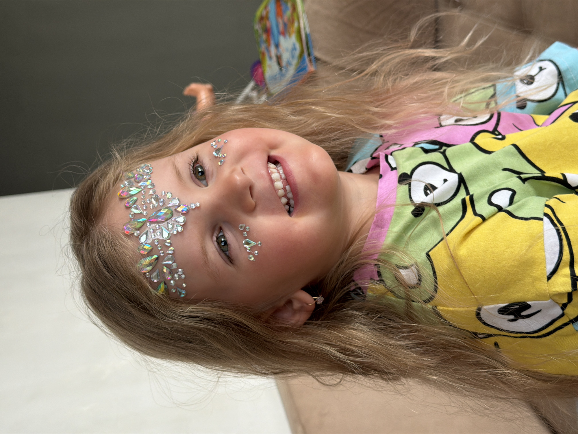 A vibrant, slightly Dutch-angled, medium close-up photograph captures a joyful young child, likely a girl, in what appears to be an indoor setting in Chaikovsky, Russia. The child, with long, flowing light brown or blonde hair, is smiling broadly at the camera, revealing her teeth and conveying a sense of genuine happiness and playfulness.
Her face is intricately decorated with an elaborate array of sparkling, iridescent, gem-like stickers or face paint. These embellishments are arranged in a delicate pattern across her forehead and extend down her cheeks, catching the light and creating a dazzling effect. A small, delicate earring is visible in her right ear.
She is wearing a brightly colored garment, possibly pajamas or a casual top, adorned with repeating cartoon panda faces in various hues of yellow, light green, pink, and light blue. The background is softly blurred, showing hints of brown furniture, likely a couch, and a partially visible colorful object, possibly a toy or book, suggesting a home environment. The natural lighting indicates the photo was taken during the daytime. There is no visible text in the image. The overall impression is one of childlike wonder and cheer during a fun, dress-up, or festive occasion.