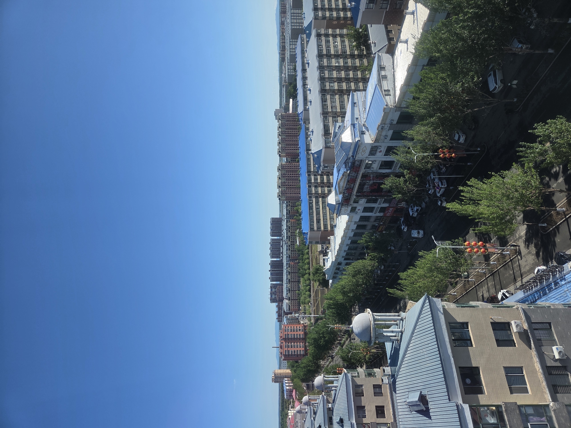 The image presents a high-angle view of an urban landscape under a clear, bright blue sky, suggesting sunny weather during daytime. The scene is dominated by numerous multi-story buildings, some with light blue sloped roofs, others with grey or brick-red facades. Green trees are abundant, lining streets and interspersed between buildings throughout the city.
In the foreground, the rooftops of several structures are visible, including one with a distinctive light blue ridged roof and a grey dome-like feature on its top. Air conditioning units are visible on the sides of buildings. Below, streets are visible with several cars, mostly sedans, parked along the curbs. A set of traffic lights with illuminated orange signals can be seen over one of the streets.
The city extends into the distance, with more densely packed buildings visible. A narrow body of water, likely a river, cuts across the horizon line, separating the foreground city from further urban development on the opposite bank. The overall impression is that of a modern, well-developed city. No discernible human activity is clearly visible, emphasizing the architectural and natural elements of the urban environment. Faint, indistinct Chinese characters appear on a sign on a building in the lower part of the image, but are unreadable.
