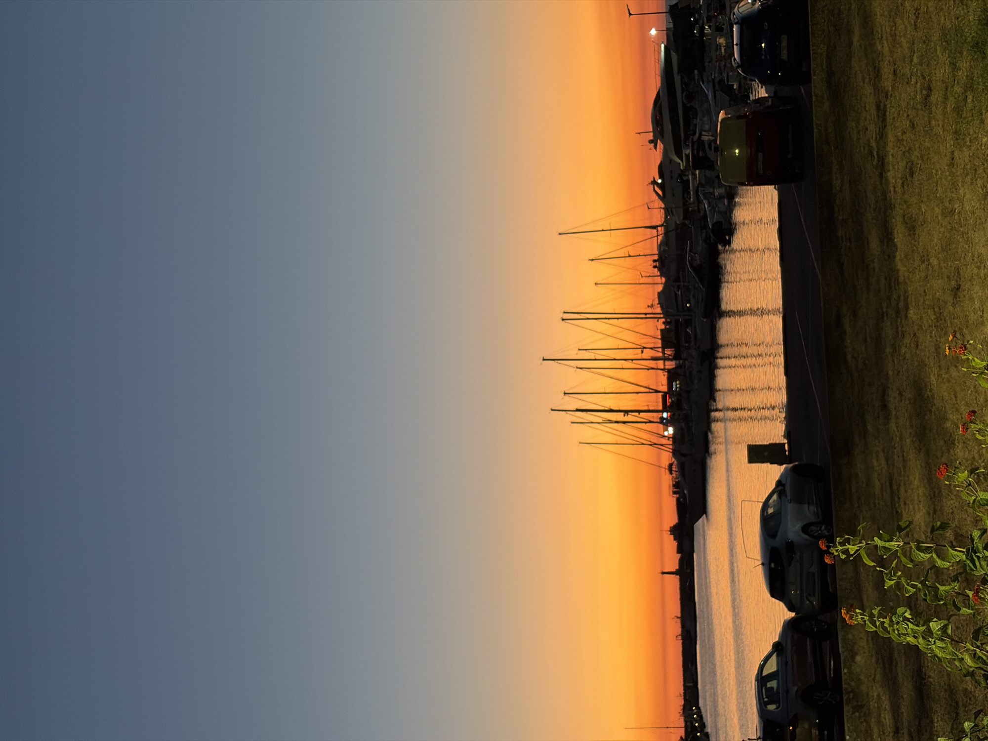 The image depicts a waterfront scene at sunset in the city of Bar, Montenegro. The sky transitions from a clear, pale blue at the top to vibrant shades of orange and yellow near the horizon. A marina filled with numerous sailboats and yachts is visible in the mid-ground, their masts silhouetted against the colorful sky. The water reflects the warm hues of the sunset. In the foreground, a grassy area is present, with some flowering plants. Several parked cars, including what appears to be a red and yellow van, are visible along the waterfront promenade. A distant lighthouse can be seen on the left side of the marina. The overall atmosphere is serene and tranquil, characteristic of a coastal sunset.