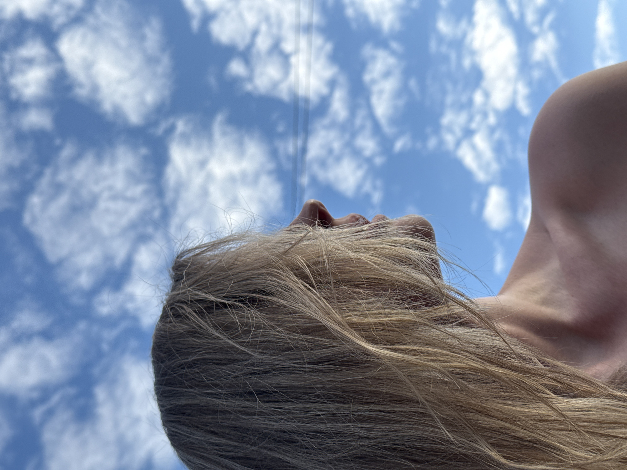 A low-angle shot reveals a person with fair skin and light brown/blonde hair, likely lying down, with their head tilted back to face the sky. The nose and lips are visible. The subject's neck and shoulder are in the right-hand portion of the image, while the hair occupies most of the bottom half. The background features a bright blue sky filled with scattered white clouds. Several vertical lines are visible in the top portion of the frame. The location and context are unspecified, so the exact location in Chaikovsky, Russia, cannot be determined. The image depicts a human looking up at the sky. No specific actions or interactions are observable.