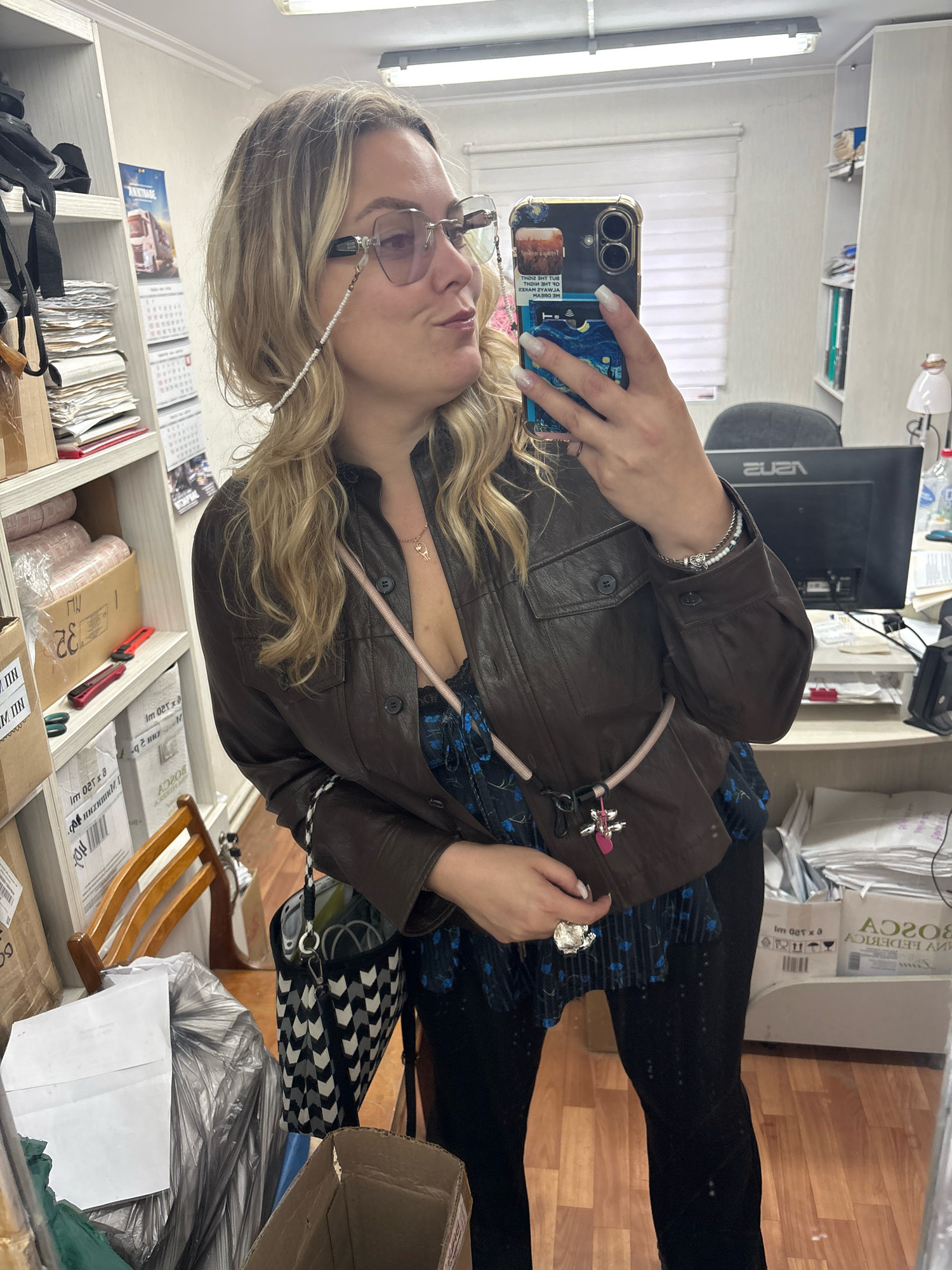 A blonde woman is taking a selfie in a cluttered office space. She is wearing glasses with a beaded chain, a brown leather jacket over a blue floral top, and black pants. She is also wearing a bracelet and has a purse hanging from her shoulder. The office is filled with shelves of boxes, paper, and office supplies. There is a computer monitor and chair visible in the background. The scene appears to be an office in Noviy, Russia, based on the presence of Russian writing on some of the boxes.
