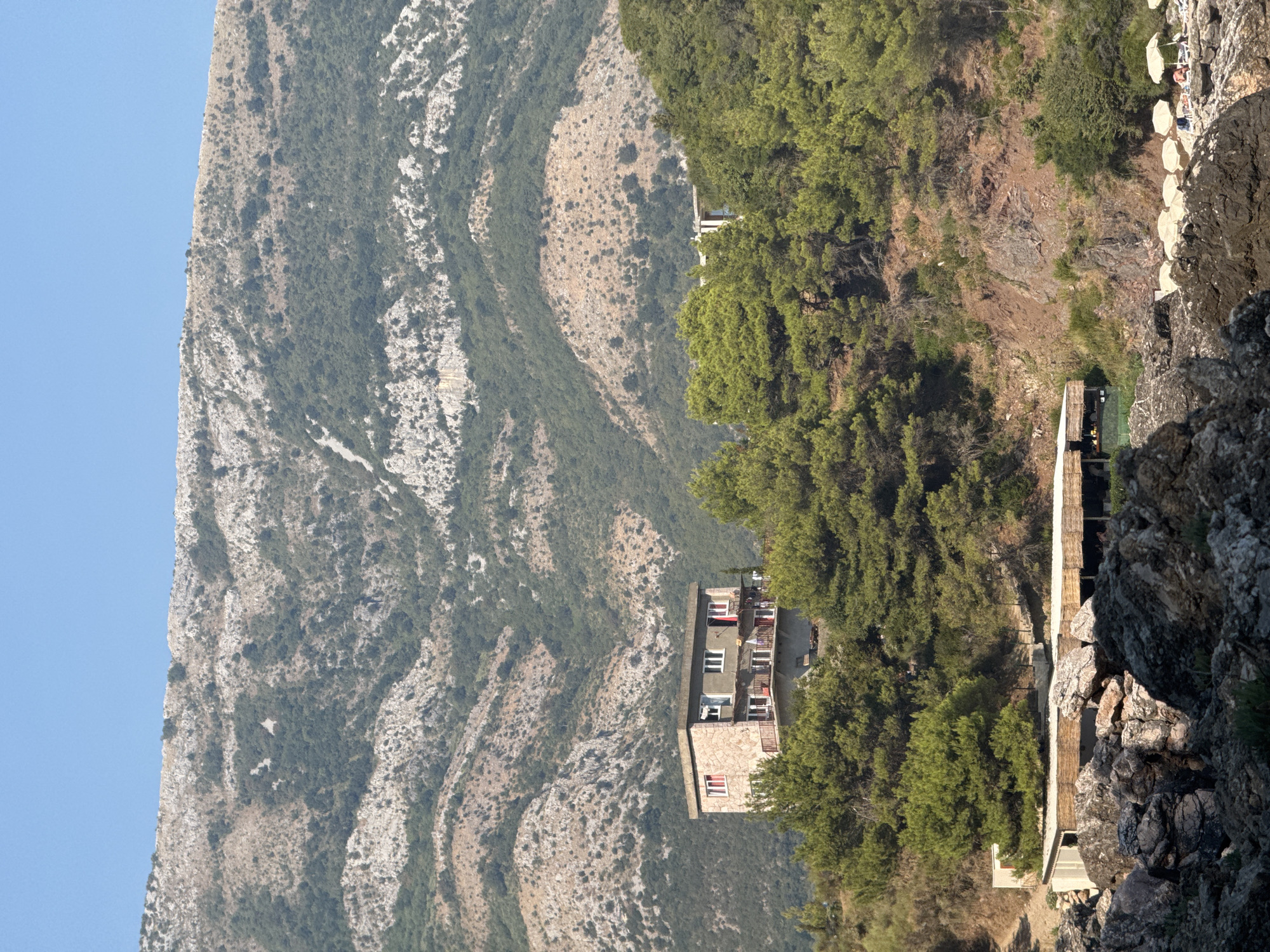 The image depicts a rocky coastline in Sutomore, Montenegro, under a clear blue sky. A multi-story building with a stone facade and red-trimmed windows sits on a slope amidst a dense forest of green pine trees. In the foreground, large, weathered rocks line the shore, with a wooden structure and umbrellas visible in the distance. The mountainous terrain, covered in scrub and trees, forms the backdrop to the scene.