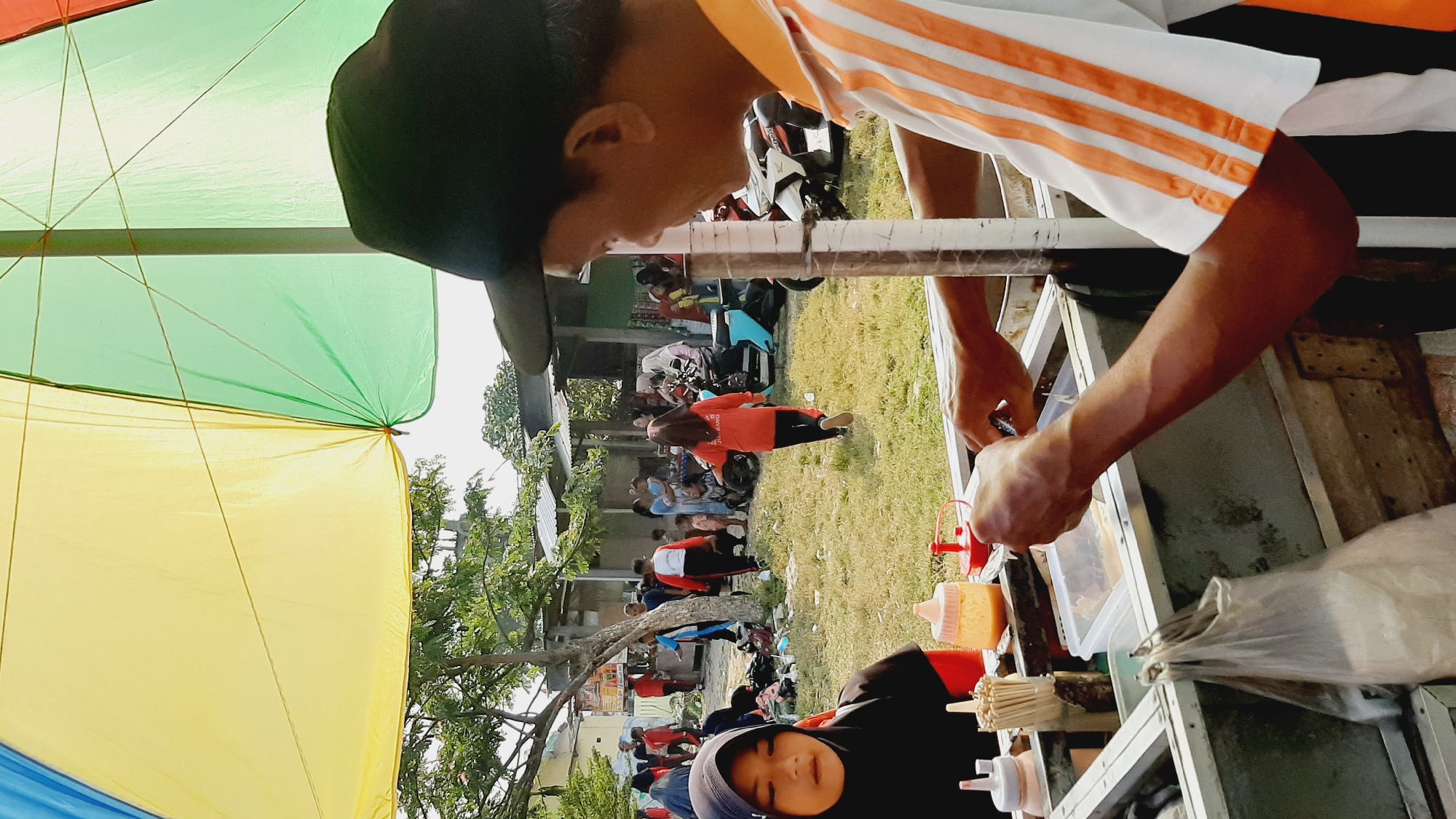 In the city of Sumberjo, Indonesia, an outdoor scene unfolds under a large, multi-colored umbrella, casting a vibrant yellow and green hue. A vendor, wearing an orange and white striped shirt and a black baseball cap, is actively preparing food. His hands are engaged in a task involving a clear plastic container, suggesting he's assembling or serving a dish. In the foreground, a young girl wearing a black hijab and an orange shirt looks on, positioned near a food cart laden with condiments, skewers, and plastic bags. The background reveals a bustling environment with numerous people, some walking, others gathered, and several motorbikes parked nearby, indicating a lively public space, possibly a market or a community event. The ground is covered in dry grass, and simple structures with corrugated metal roofs are visible in the distance, characteristic of the local architecture. The atmosphere appears to be daytime, with bright sunlight.